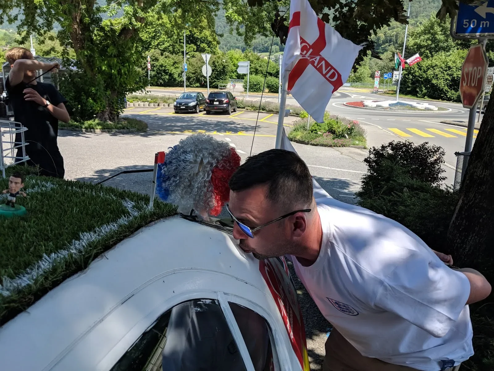 A man with sunglasses appears to be kissing the hood of a small car decorated with grass and a figurine, during an outdoor event with flags, trees, and a parking lot in the background.