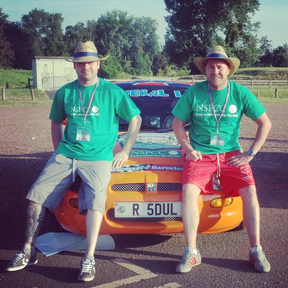 Two men wearing green NSPCC shirts and straw hats, standing on either side of a bright orange race car, with a background of trees and a parking lot.
