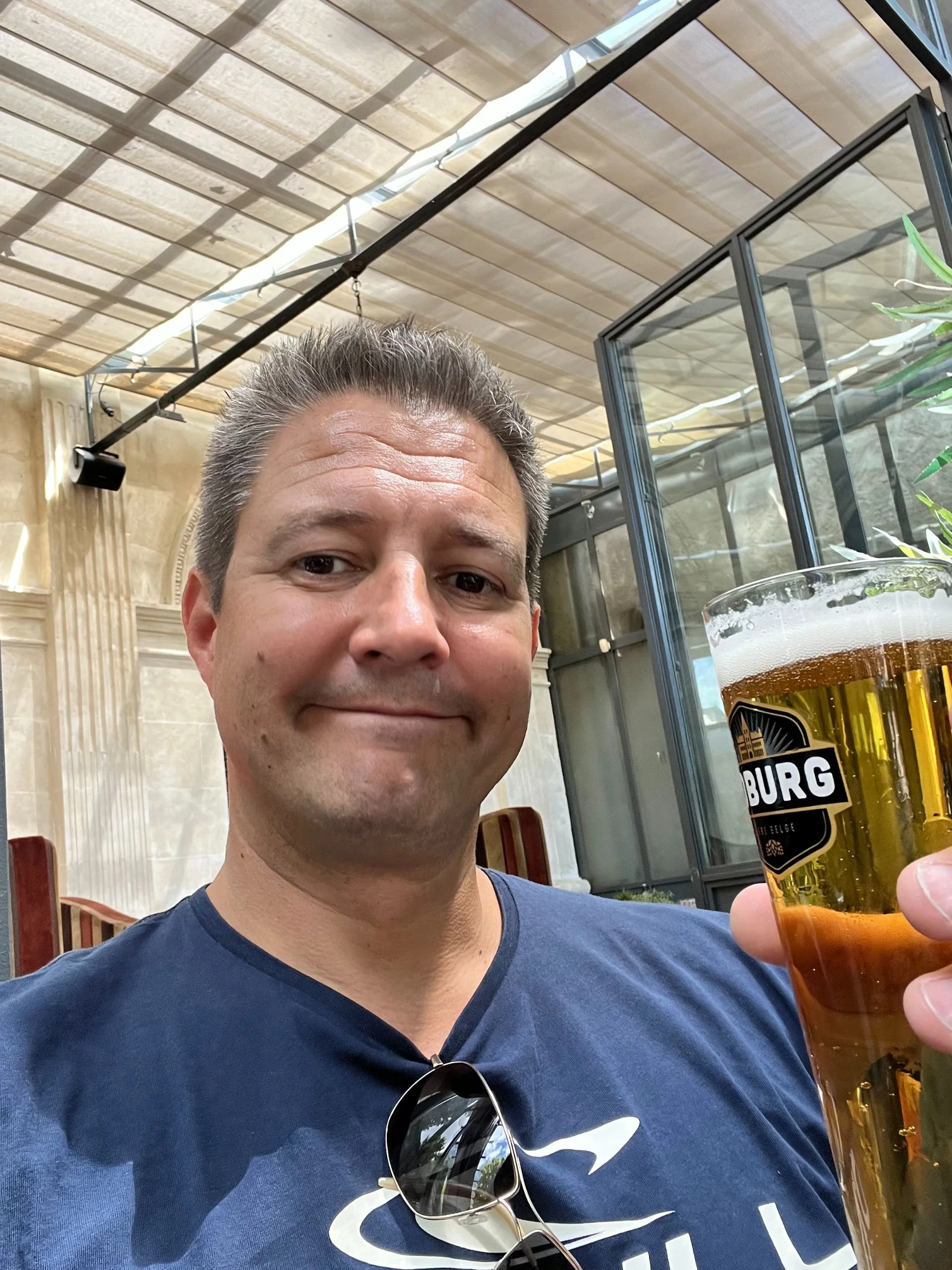 A man in a blue shirt holding a pint of beer at an indoor venue with glass walls and a covered outdoor area.