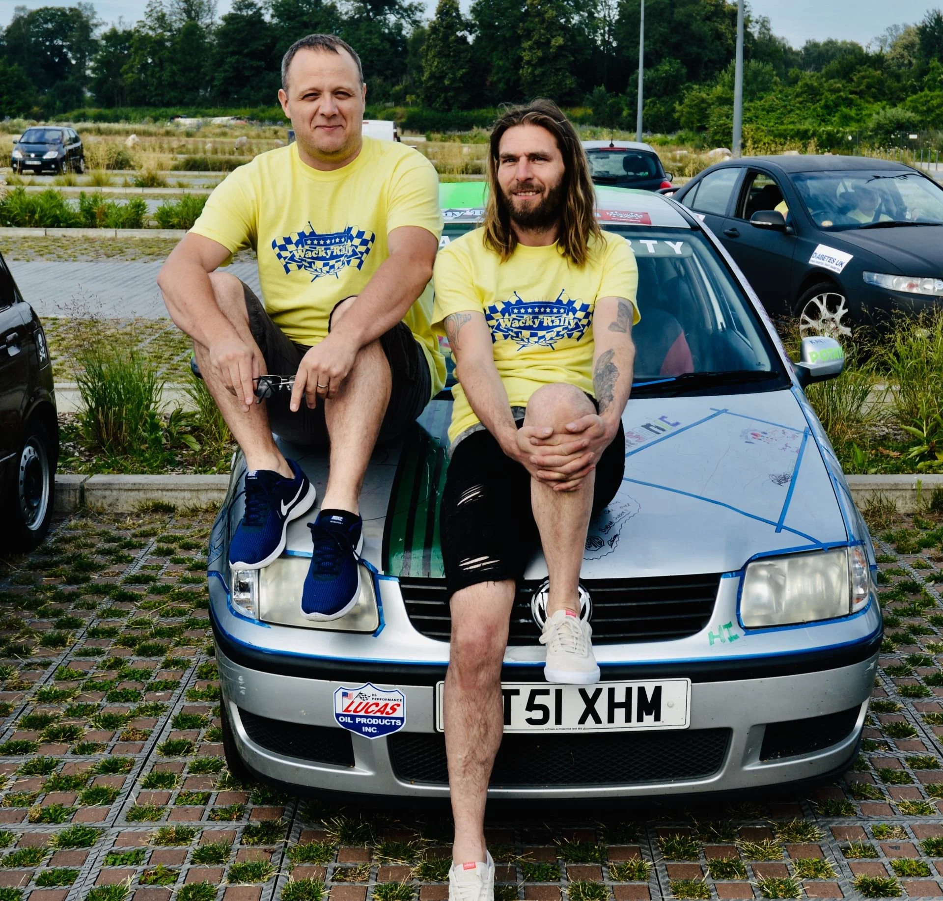 Two men in yellow T-shirts with 'Wacky Rally' logo sitting on the hood of a gray race car in a parking lot, with other cars and greenery in the background.