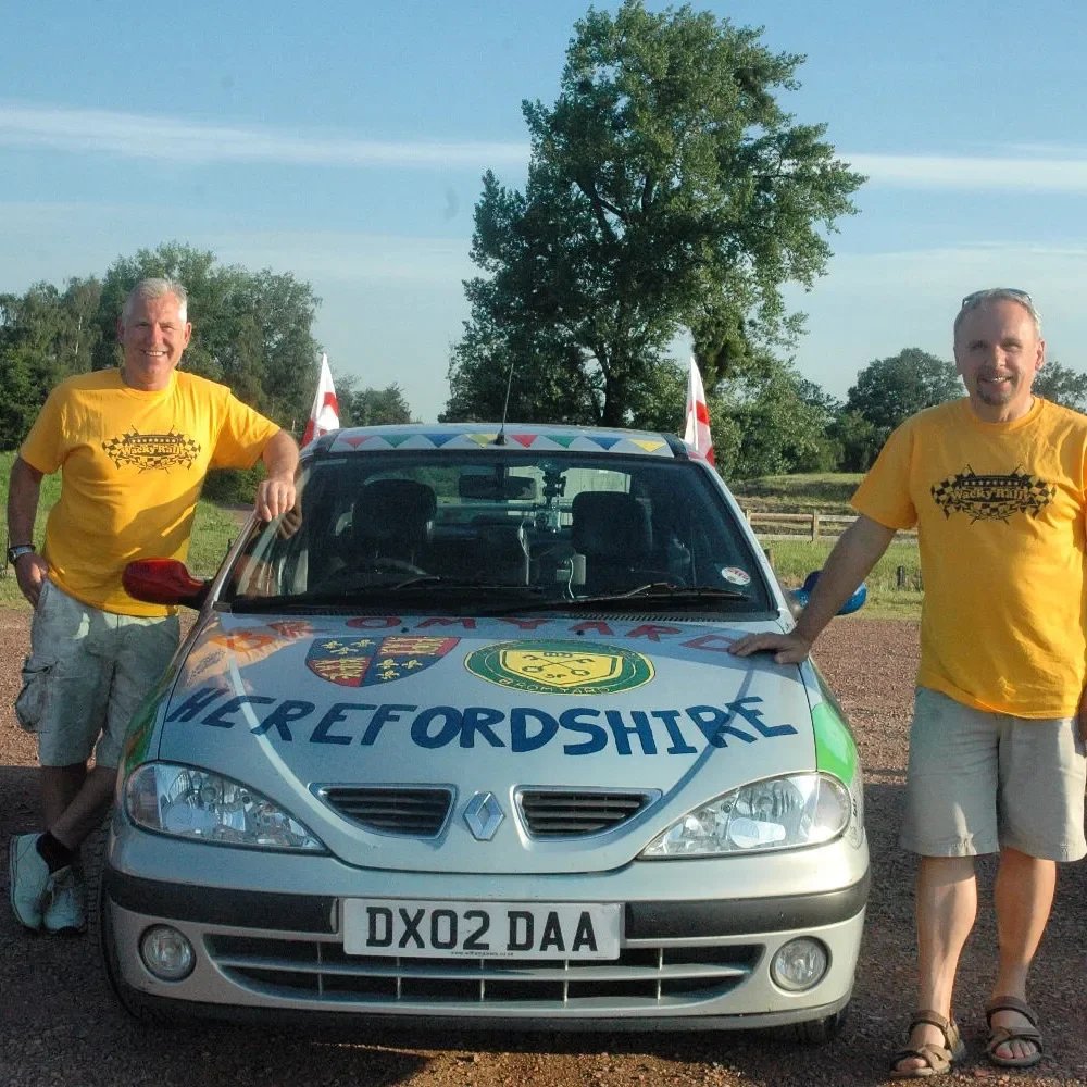 Two men standing next to a decorated car with Hertfordshire writing and symbols, on a gravel area with trees and a clear sky in the background, both wearing yellow shirts and smiling.