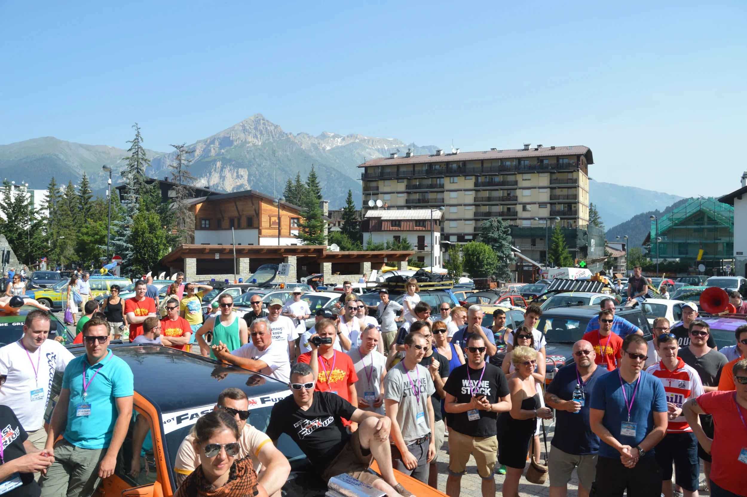 Large crowd of people gathered outdoors in front of various parked cars, with mountains and trees in the background on a sunny day.