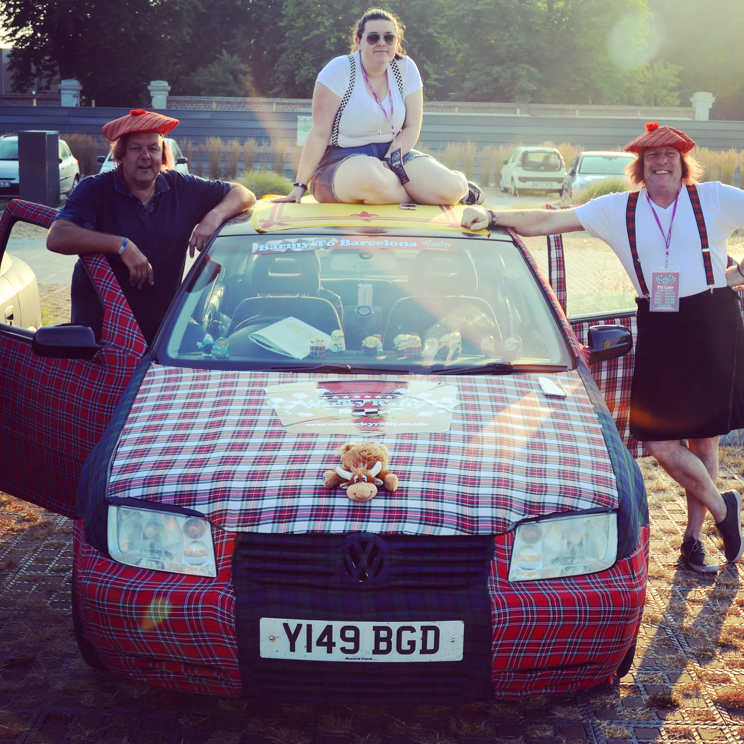 People posing with a car decorated in plaid fabric, with two men wearing matching plaid hats, a woman sitting on top of the car, and a plush moose toy on the hood of the car. The car has a sign on the front that says 'Y149 BGD' and a sticker that say