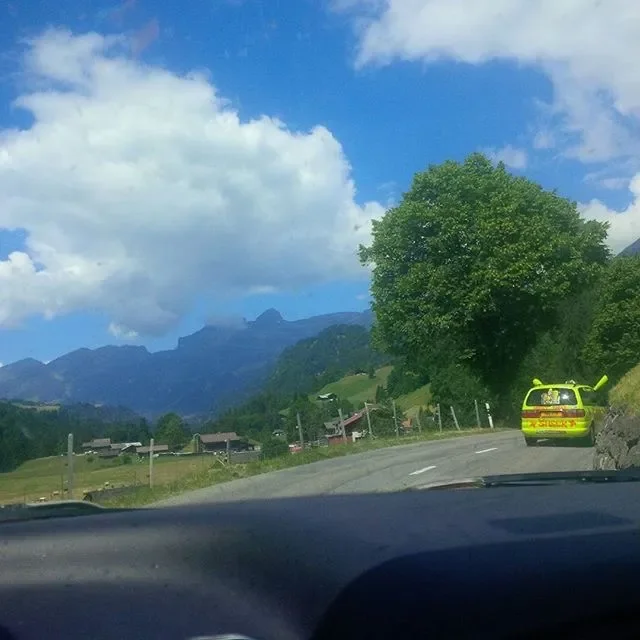 A roadside scene with a green Volkswagen Golf with a snorkel, parked on the shoulder next to a large tree. In the background are hills or mountains under a partly cloudy blue sky.