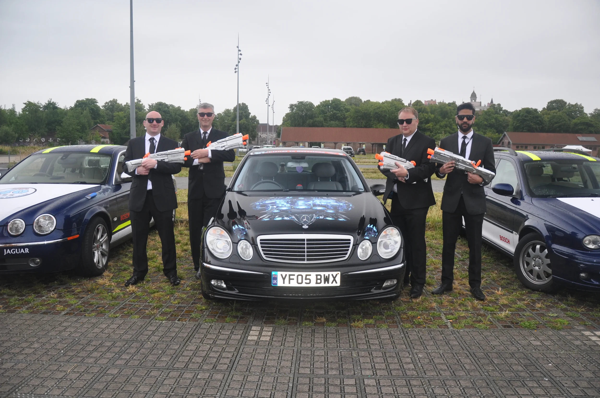 Four men in black suits and sunglasses standing in front of black Mercedes-Benz with a car logo, holding large toy guns, flanked by blue cars with racing decals, on a parking lot with overcast sky.