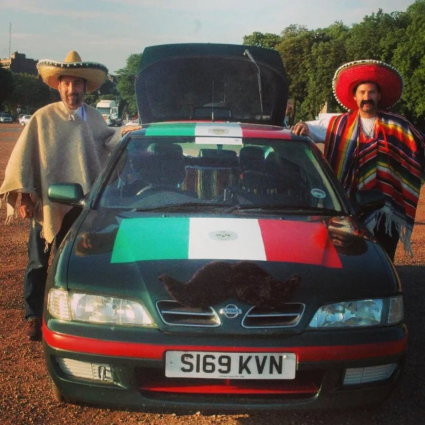 Two men wearing traditional Mexican outfits with sombreros standing beside a Nissan car decorated with Mexico's flag colors. The car has a black cat lying on the hood and an open trunk, with a national flag on top of the trunk. The background shows t