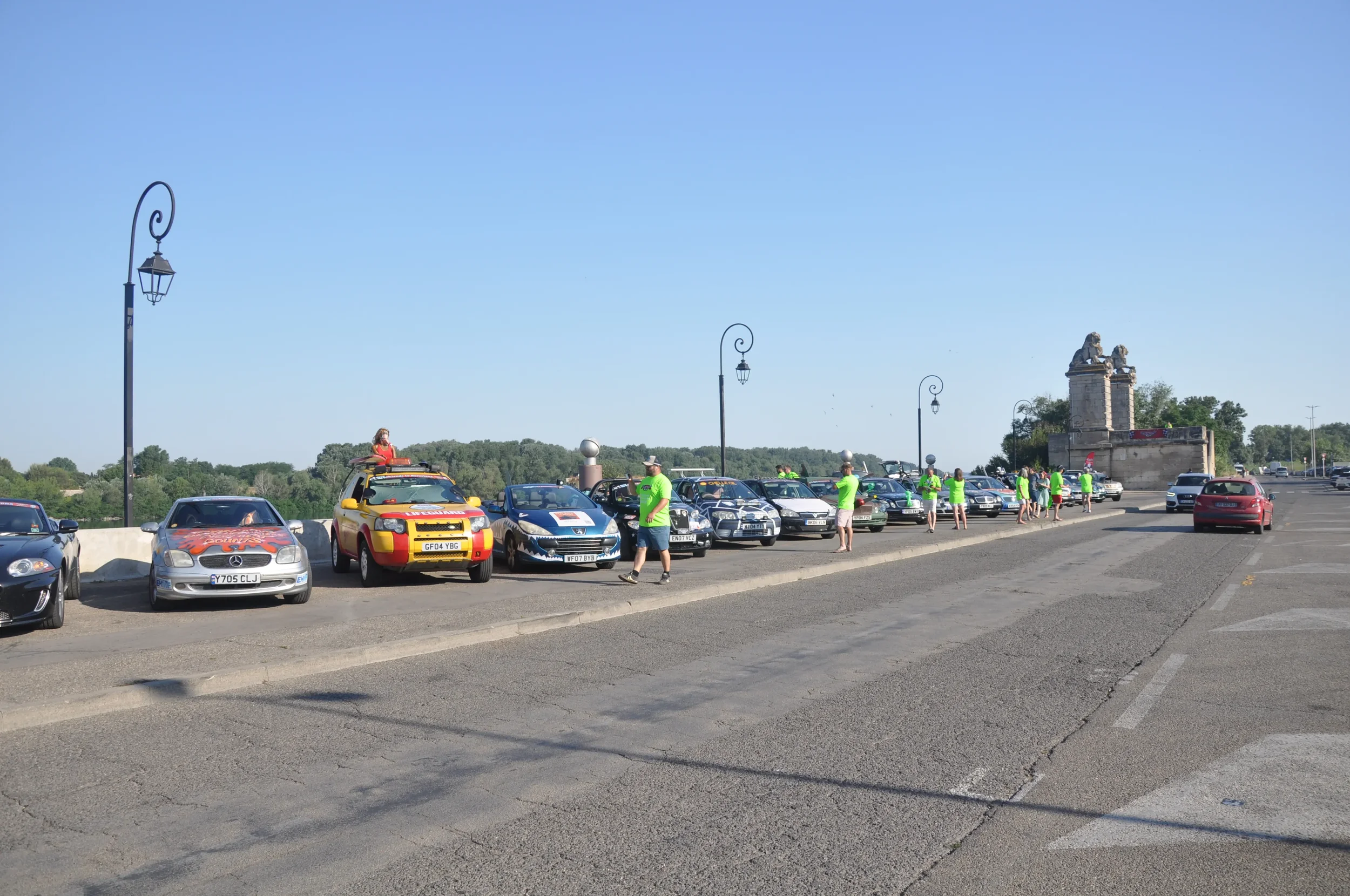 Group of people in neon green shirts standing near parked cars along a bridge with tall lampposts and a stone monument in the background, on a sunny day.