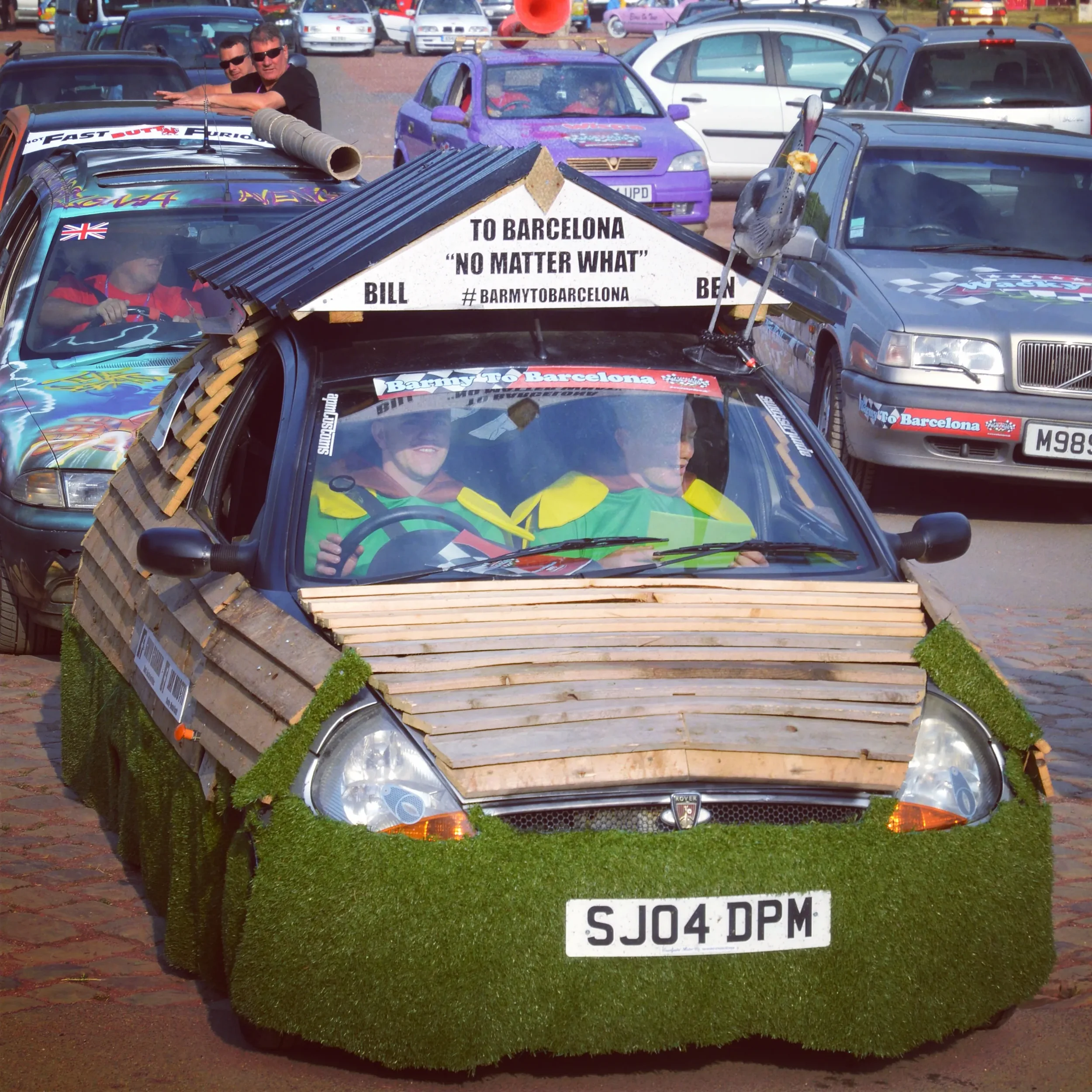 A small car decorated with wooden planks, grass, and other materials to look like a themed craft. The car has a sign on top that reads "To Barcelona 'No Matter What'" and stickers referencing the event. Two people are inside the car, dressed in green