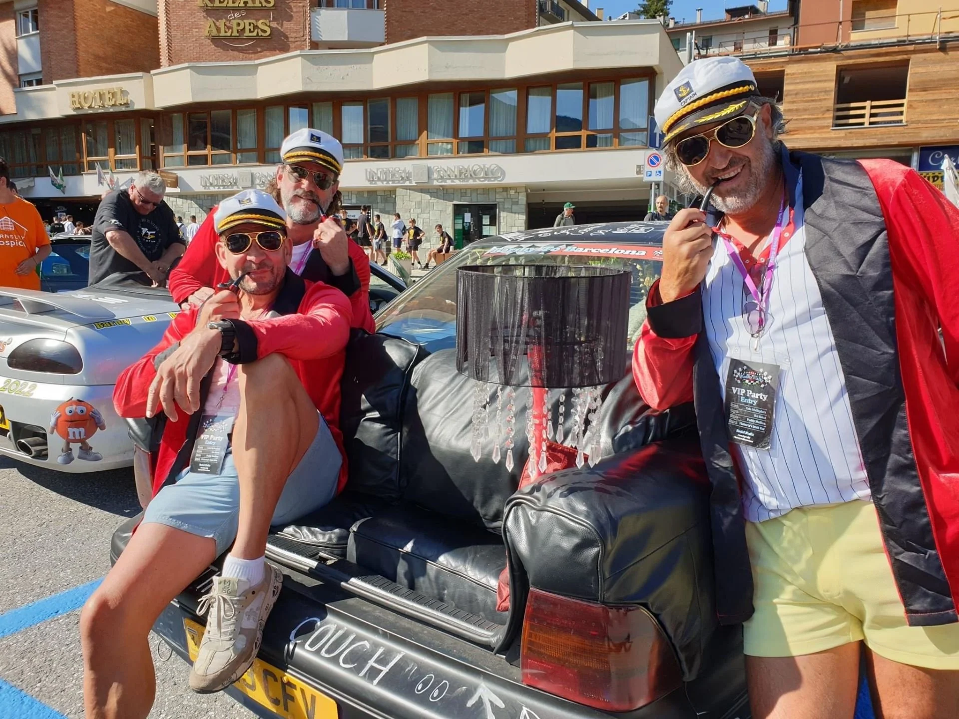 Three men dressed in racing-themed outfits standing near a car with a sign that says 'Couch' on it, in front of a hotel and a crowd of people.