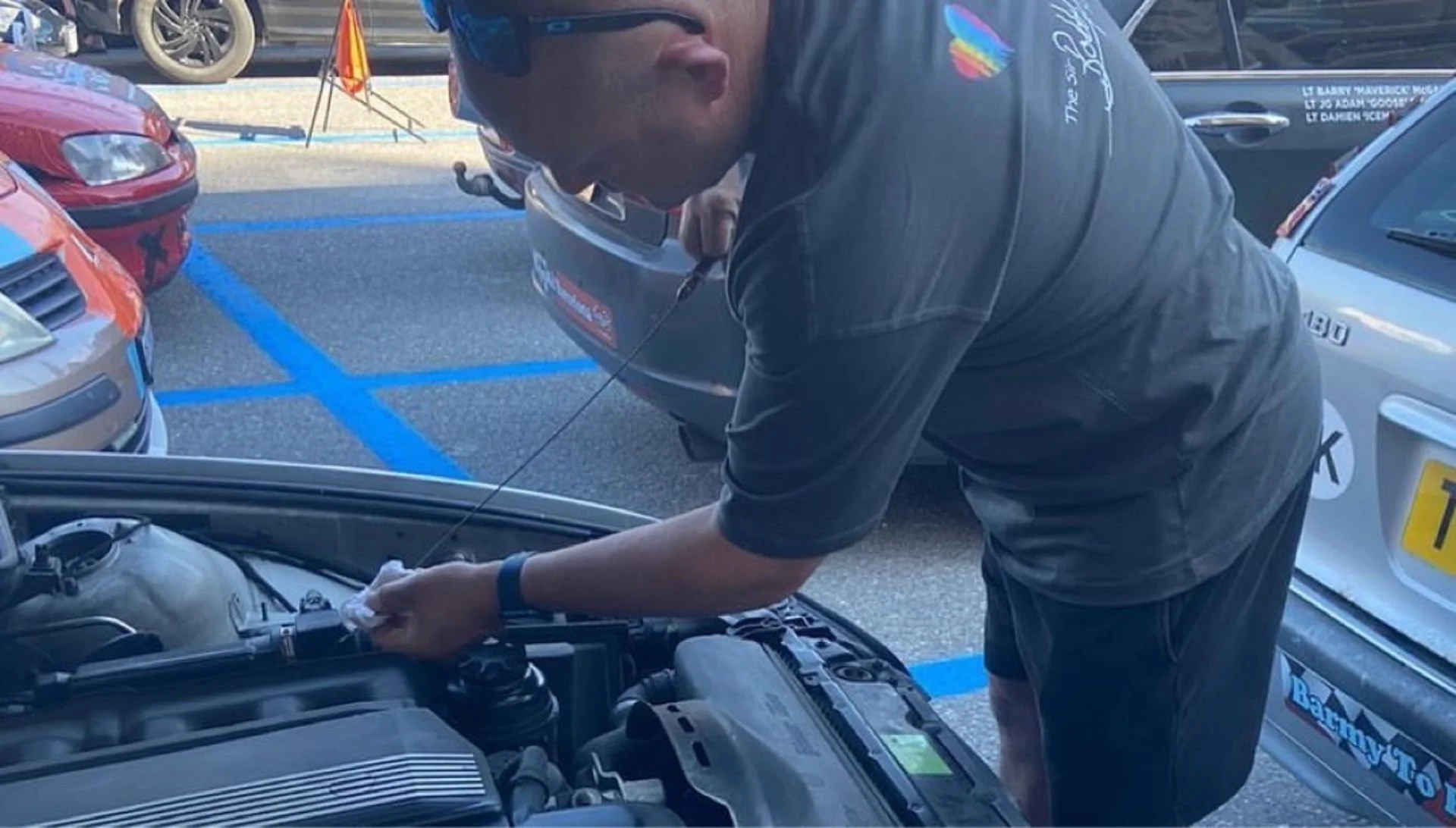 A man wearing sunglasses and a black t-shirt working on a car engine in a parking lot with other cars around.