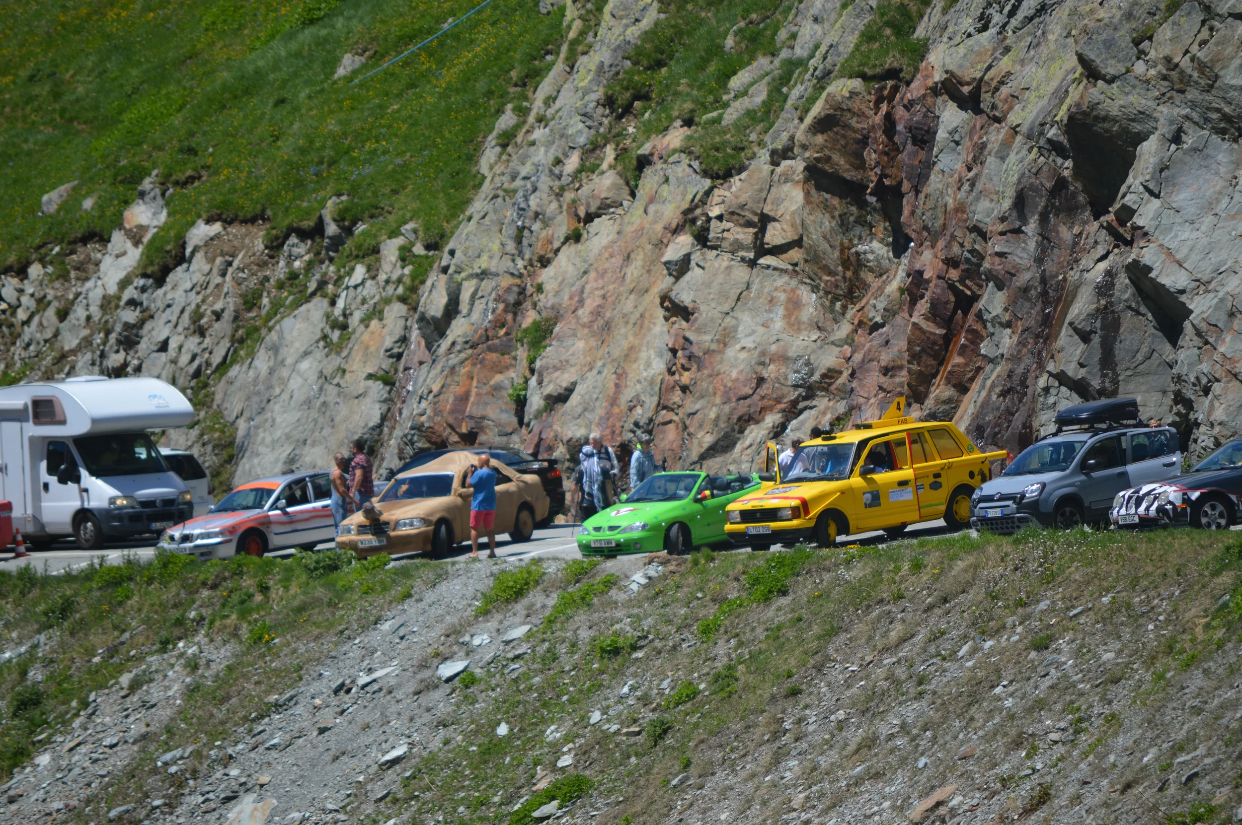 Multiple cars and people stopped on mountain road next to a rocky cliff, some cars covered with camouflage, likely for a car test or event.