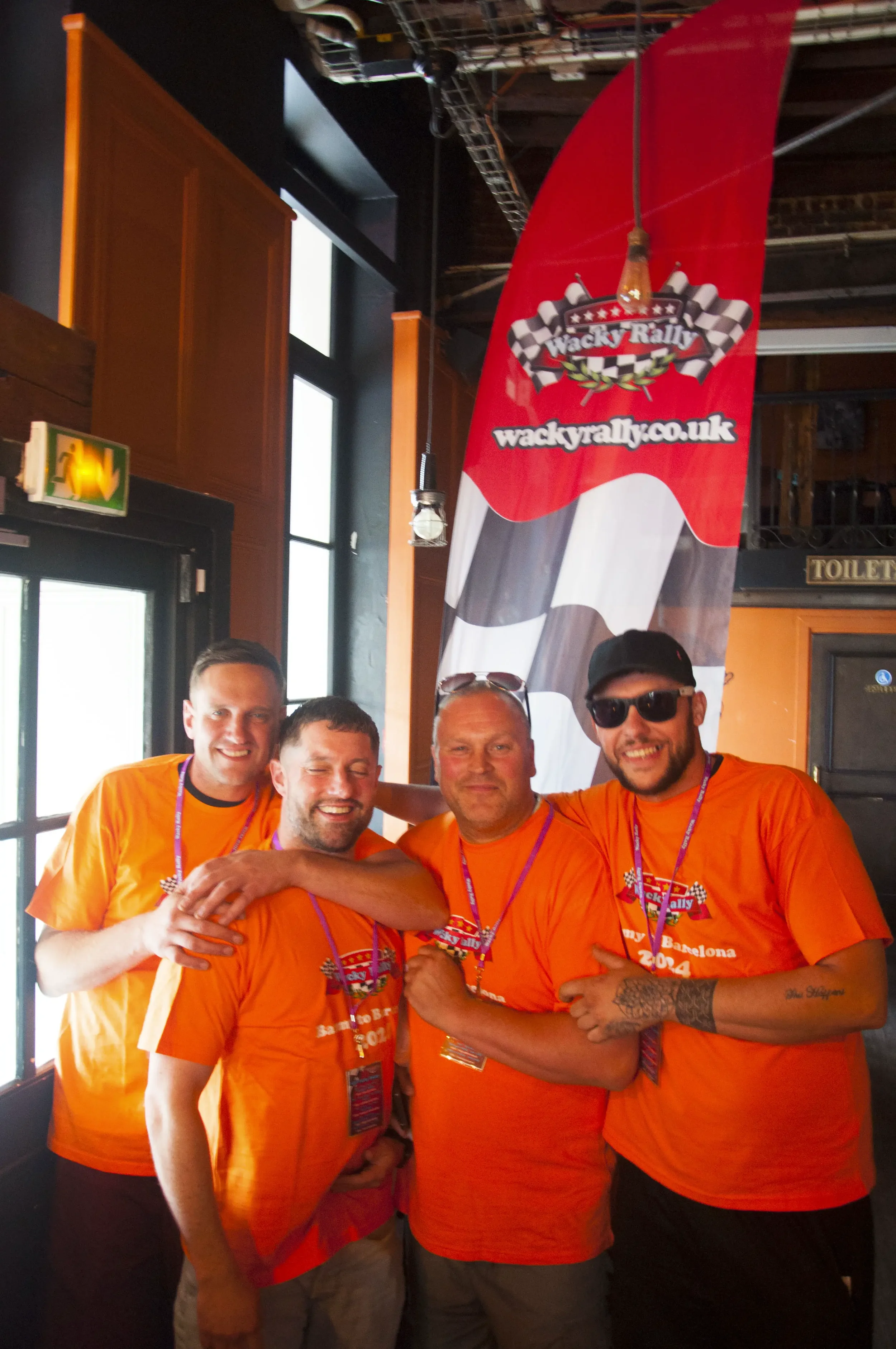 Four men in orange T-shirts celebrating at a rally event, standing in front of a red and black flag with a checkered flag design and text, inside a building with windows and a sign for toilets.