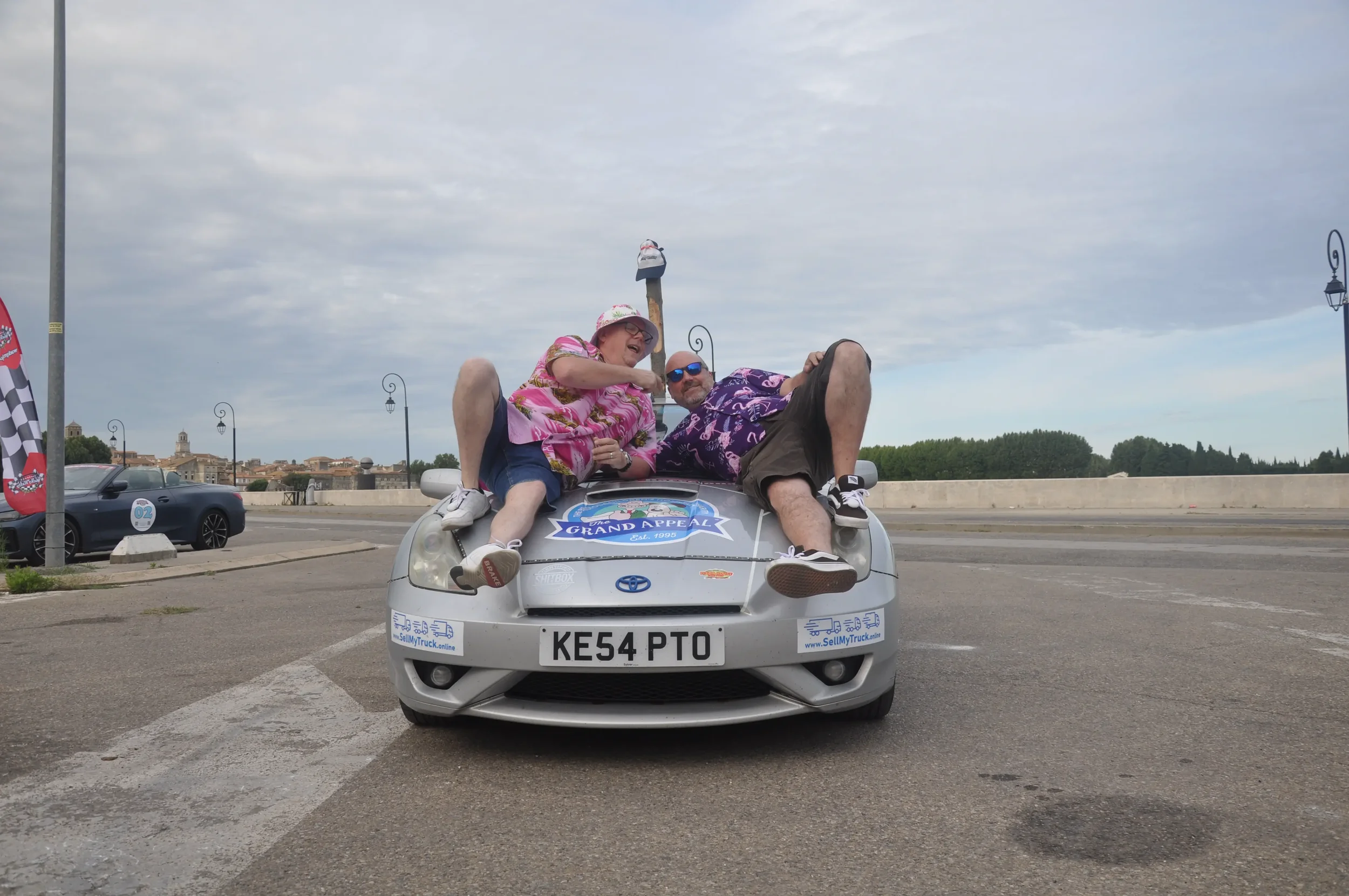 Two people dressed in colorful casual clothing relaxing on the hood of a silver Toyota car with a 'Grand Appeal' sticker, parked on a paved area with a scenic town in the background.