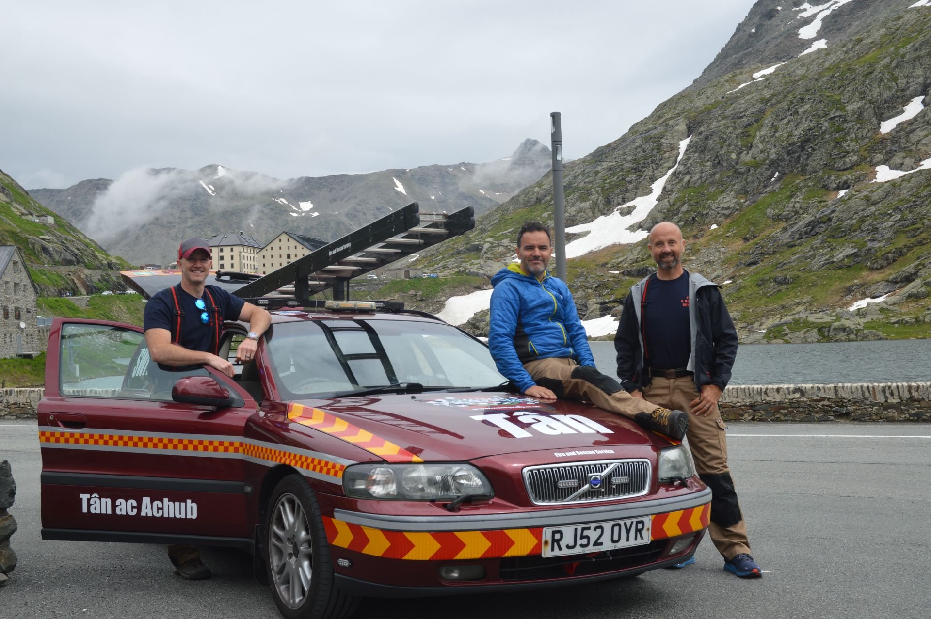 Three men standing and sitting next to a red rescue vehicle with a ladder, in a mountainous area near a lake, with snow patches on the mountains.