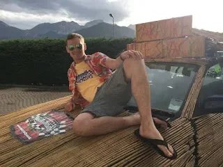 Person lounging on a car with one leg crossed over the other, wearing sunglasses, a colorful shirt, and shorts, next to a picnic blanket on a wooden platform with mountains in the background.