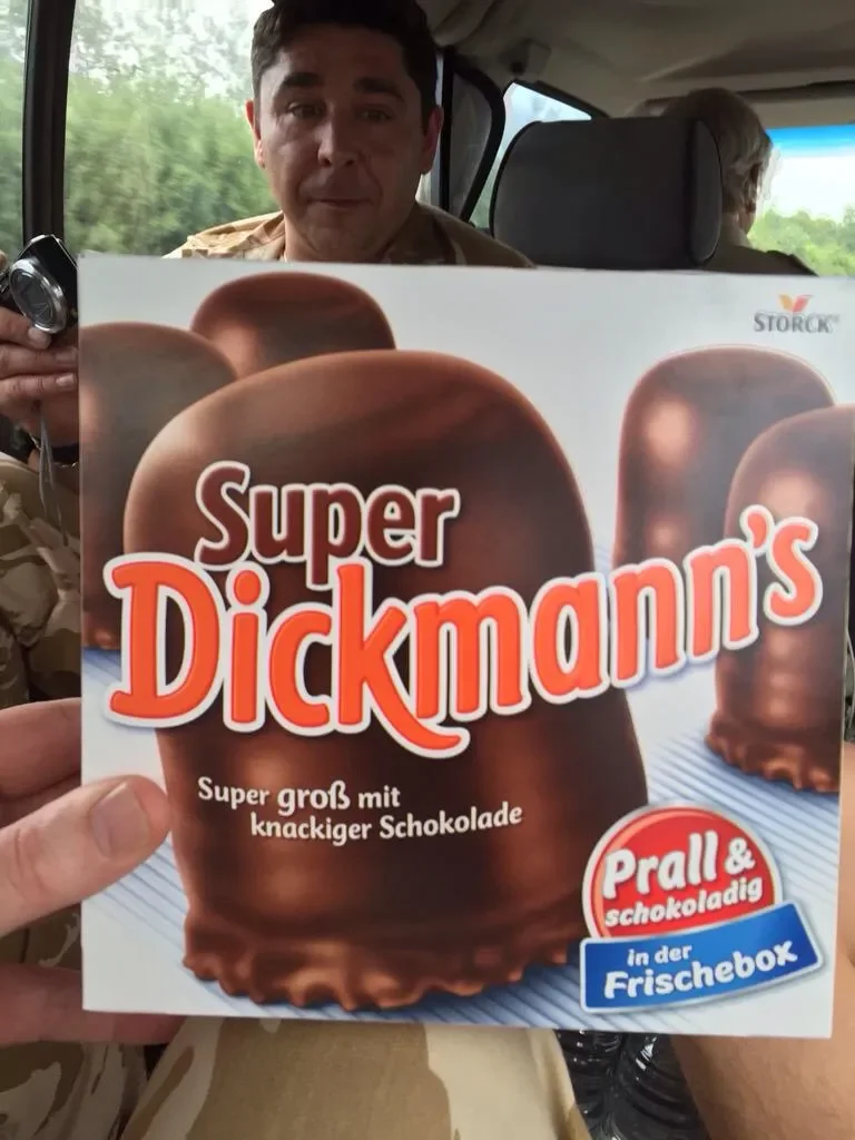 Person sitting in vehicle holding a box of Super Dickmann's ice cream bars with chocolate coating, inside a vehicle with trees visible outside the window.