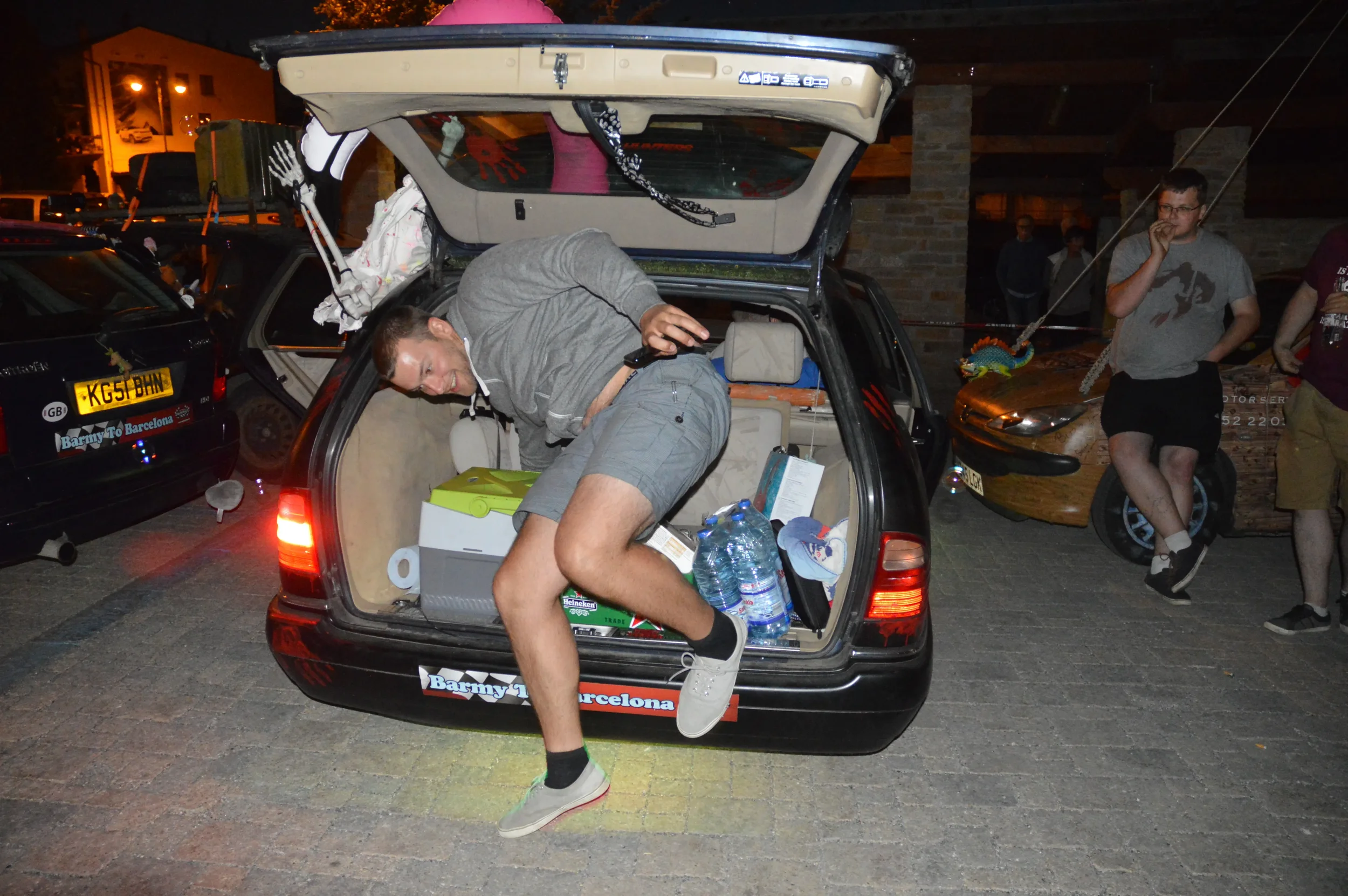 A man with a gray hoodie and shorts climbing out of the open trunk of a black car, which is filled with water bottles, boxes, and other items. There are people in the background, one leaning against a car and another standing nearby. The scene appear