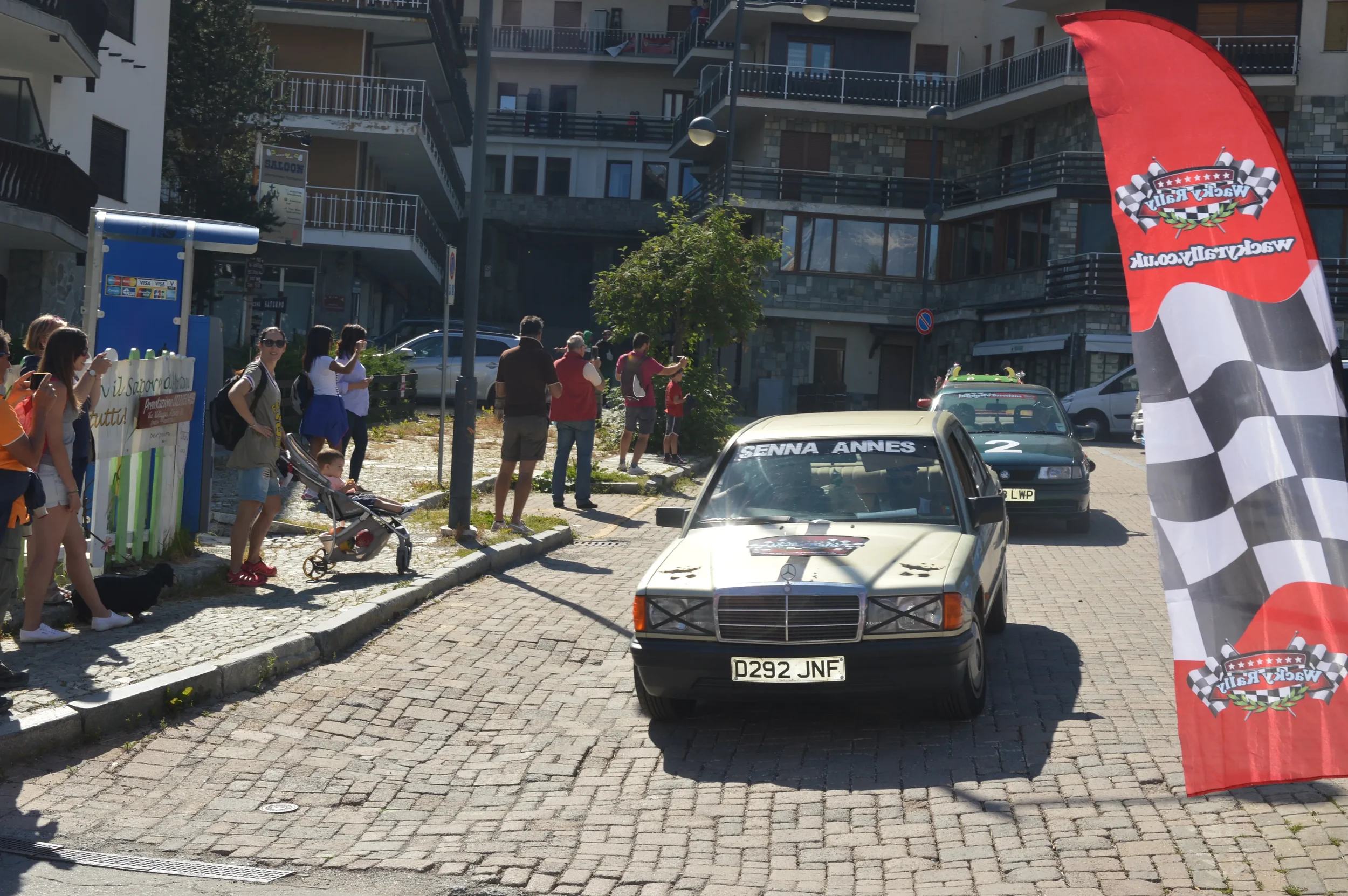 A small crowd of people on a sidewalk watching a vintage Mercedes race car in a car event on a cobblestone street, with a red checkered flag banner on the right side.
