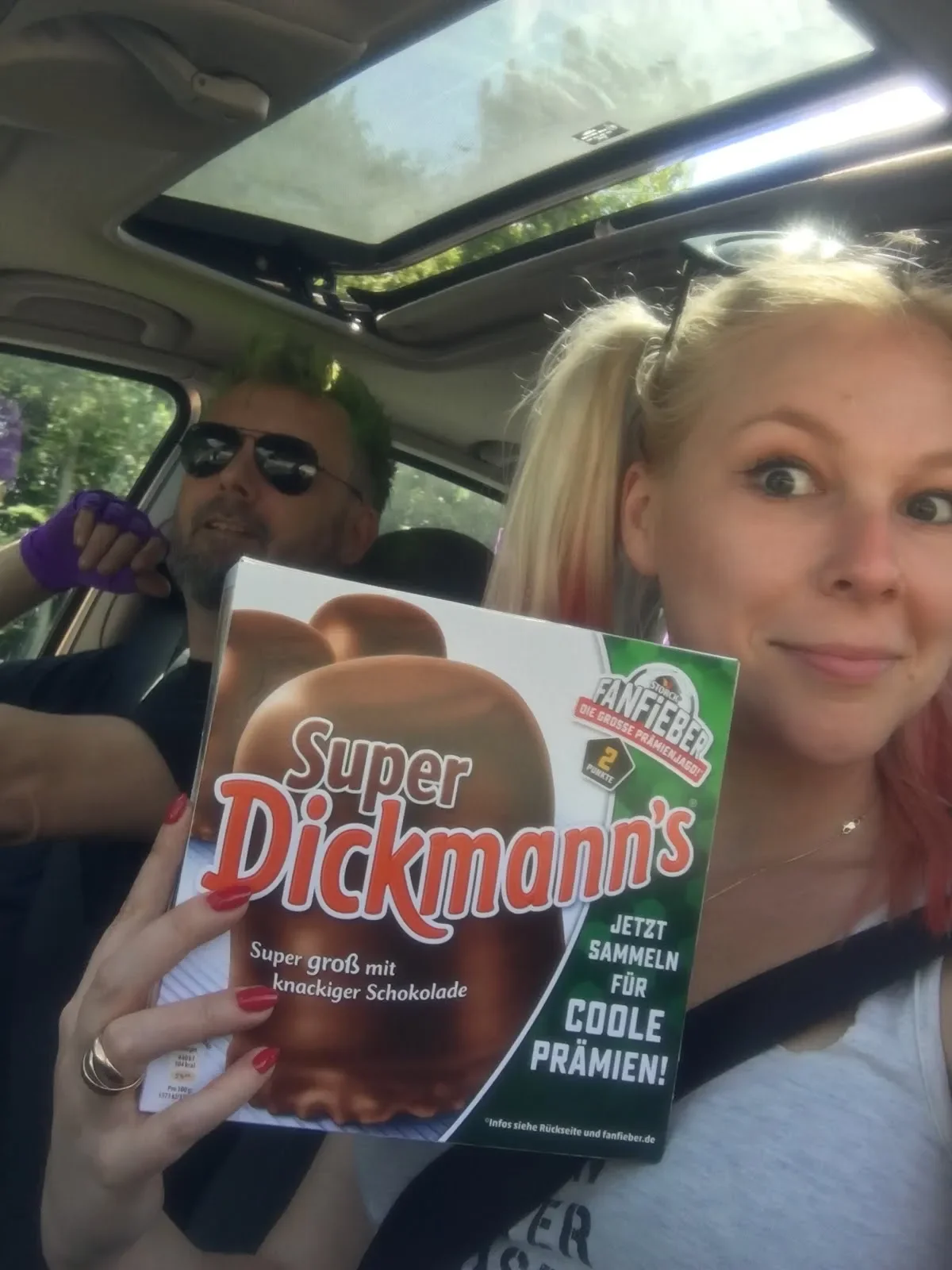A young woman with blonde hair holding a box of Super Dickmann's chocolate-covered marshmallow treats in the passenger seat of a car, with a man in sunglasses in the background.