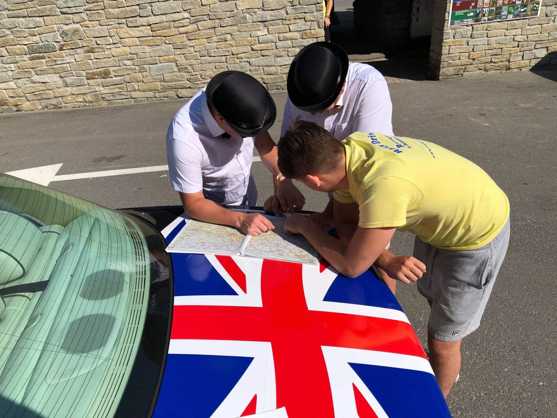 Three men are gathered around a map on the hood of a car with a Union Jack flag design. Two of the men are wearing white shirts and black bowler hats, and the third is wearing a yellow T-shirt and gray shorts. They are outdoors near a stone wall.