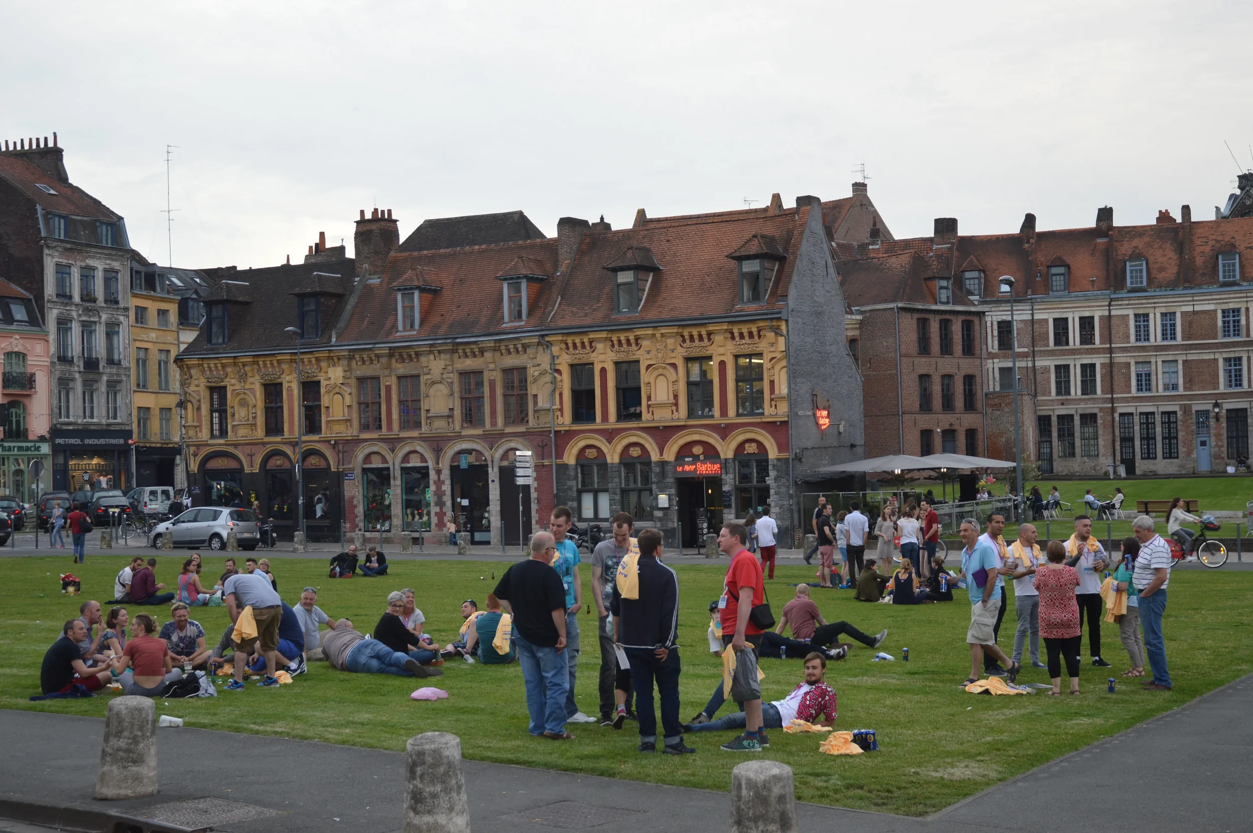People gathering and socializing on a grassy area in an urban park with historic buildings in the background during daytime.