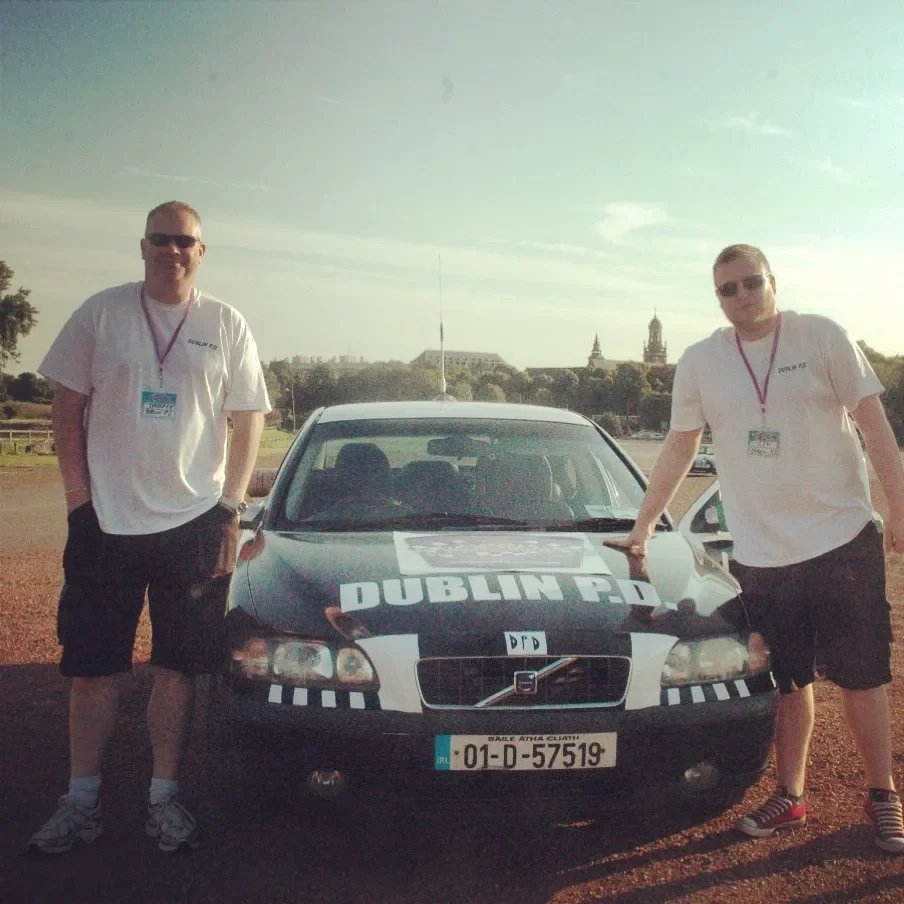 Two young men wearing sunglasses and white t-shirts with lanyards stand on either side of a black police car with 'Dublin P.D.' written on the hood. They are outdoors on a sunny day, with trees and old buildings in the background.