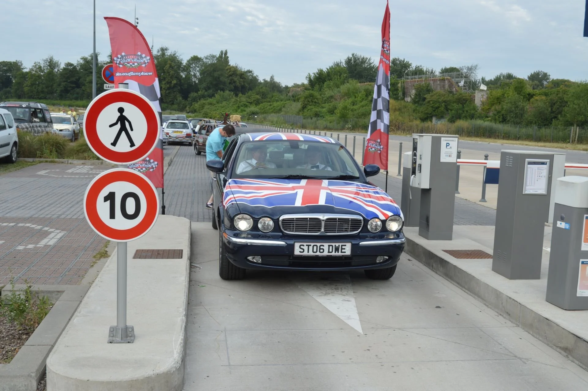 Car decorated with Union Jack flag parked at a charging station with barriers, signs, and flags around.