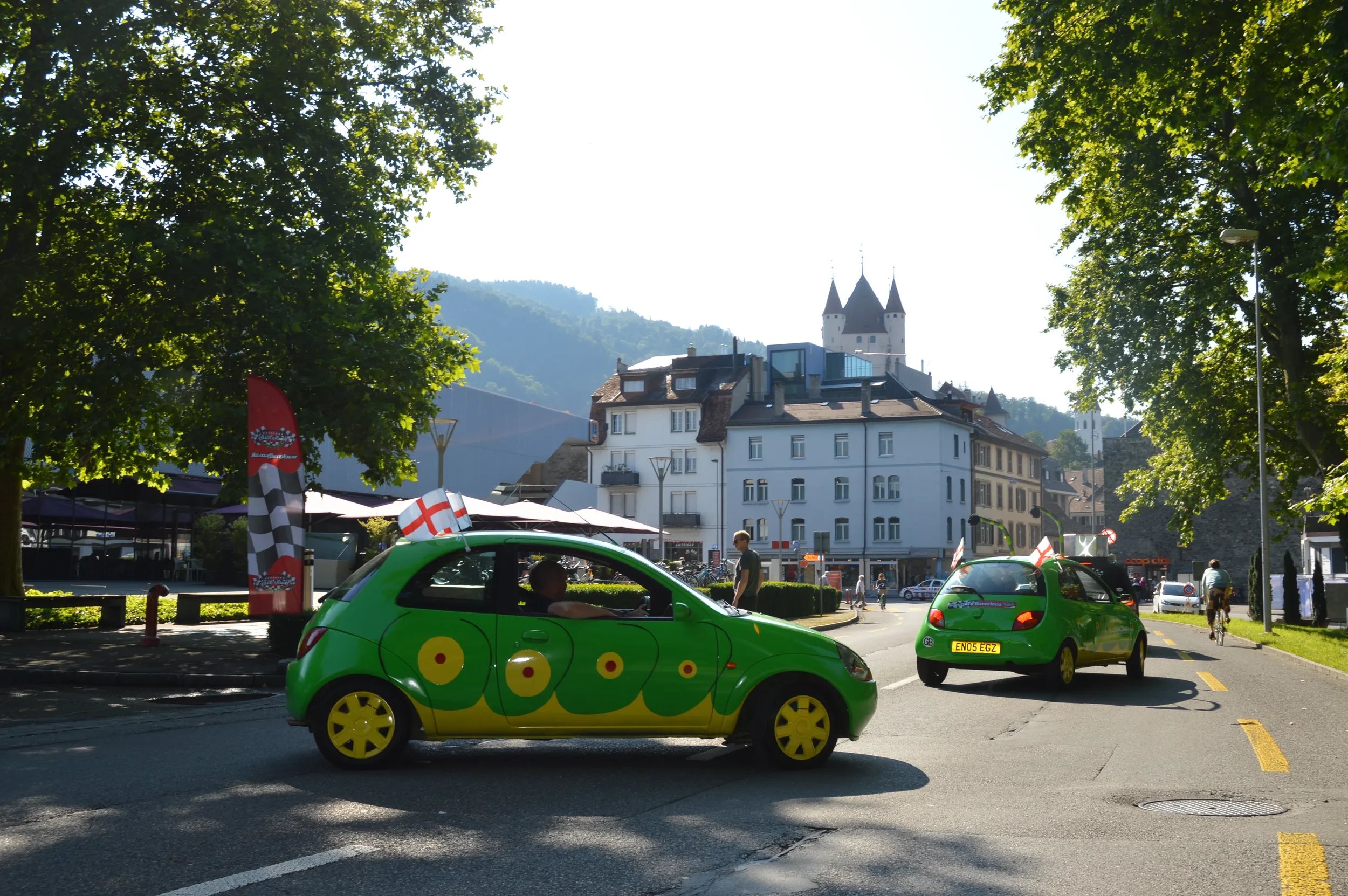 Two small green cars with yellow wheels and decorated with red, yellow, and white designs, parked in a town with trees, buildings, and a castle in the background.