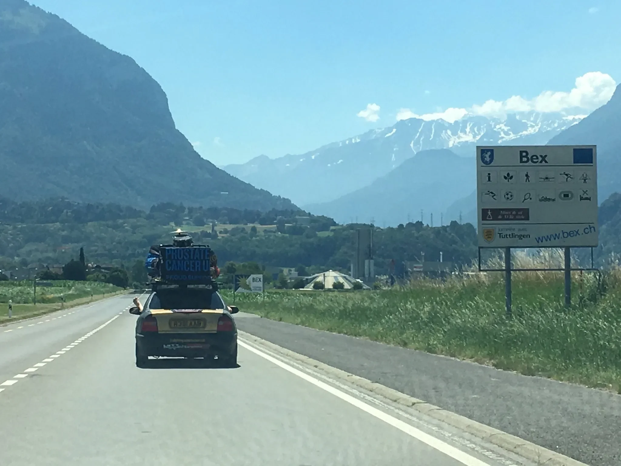 A car driving on a scenic highway with mountainous landscape in the background, bearing a sign on top that says 'PROSTATE CANCER U' and a large informational sign on the right side of the road with symbols indicating local attractions and services in