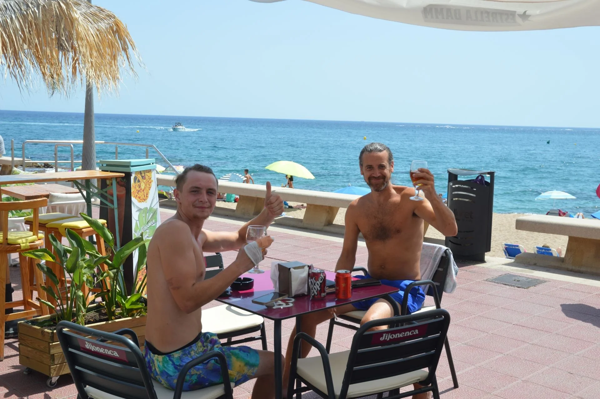 Two shirtless men sitting at a table on a beachside patio, holding drinks and smiling, with the ocean and beach umbrellas in the background.
