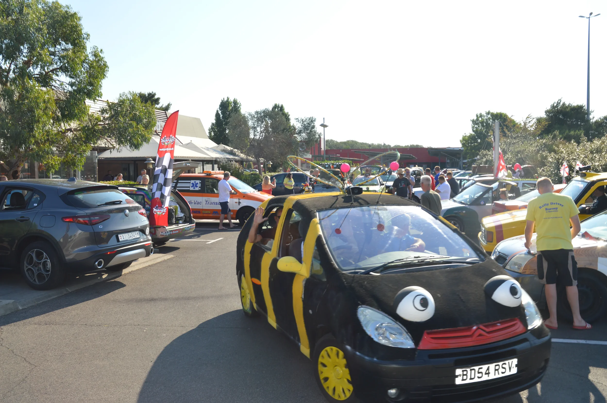 A parking lot filled with decorated cars and people at a festive outdoor event. One car resembles a black and yellow bee with cartoonish eyes, and others have various themes with flags and banners.
