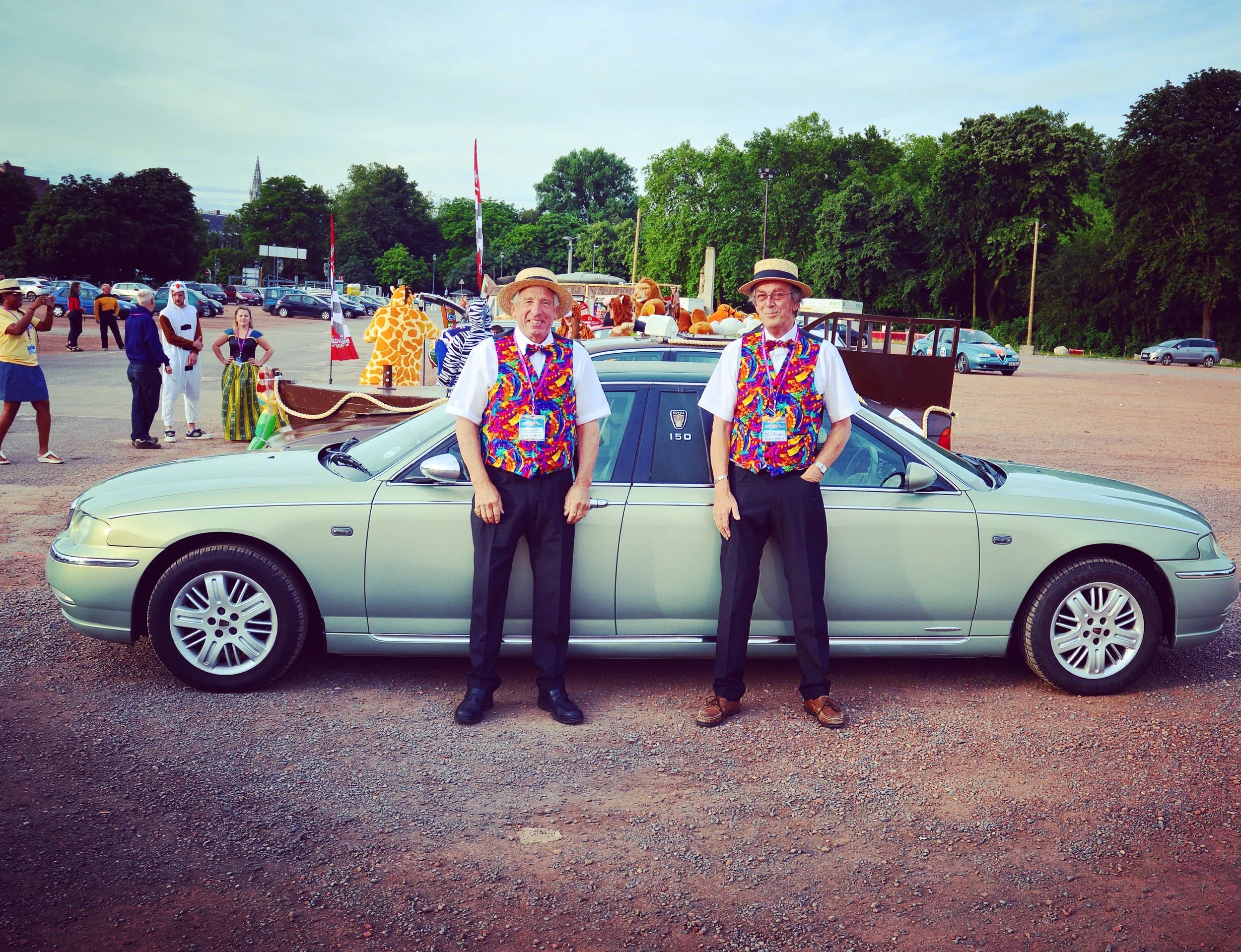 Two men dressed in colorful, patterned vests, white shirts, black pants, and straw hats standing in front of a gray car at an outdoor event with other people, animal costumes, and trees in the background.