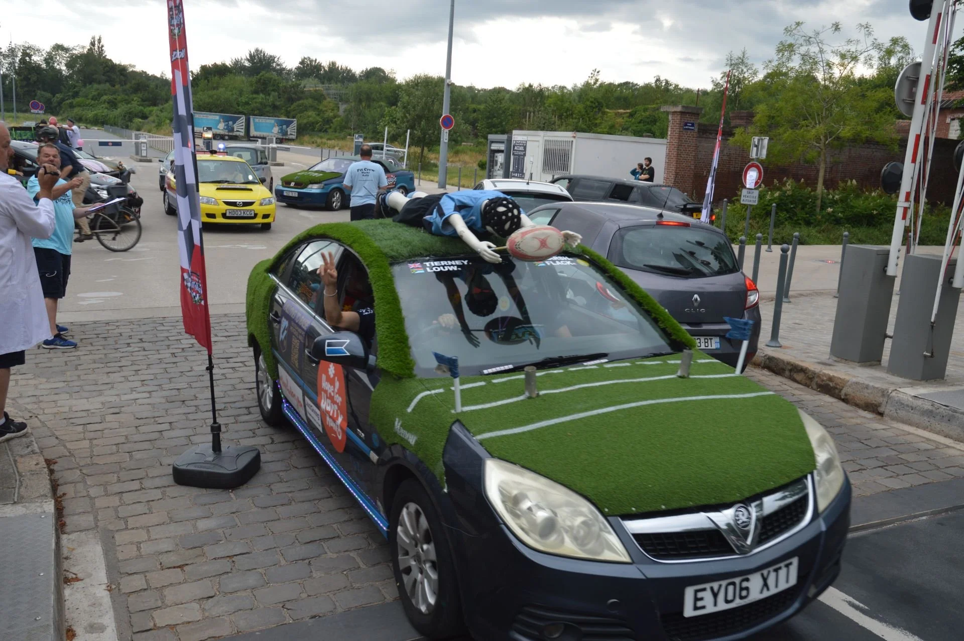 A decorated car with artificial grass on the hood and roof, and rugby goalposts on the hood, parked among other cars. A person dressed as a rugby player on top of the car is holding a rugby ball, and others are nearby at an outdoor event with people 