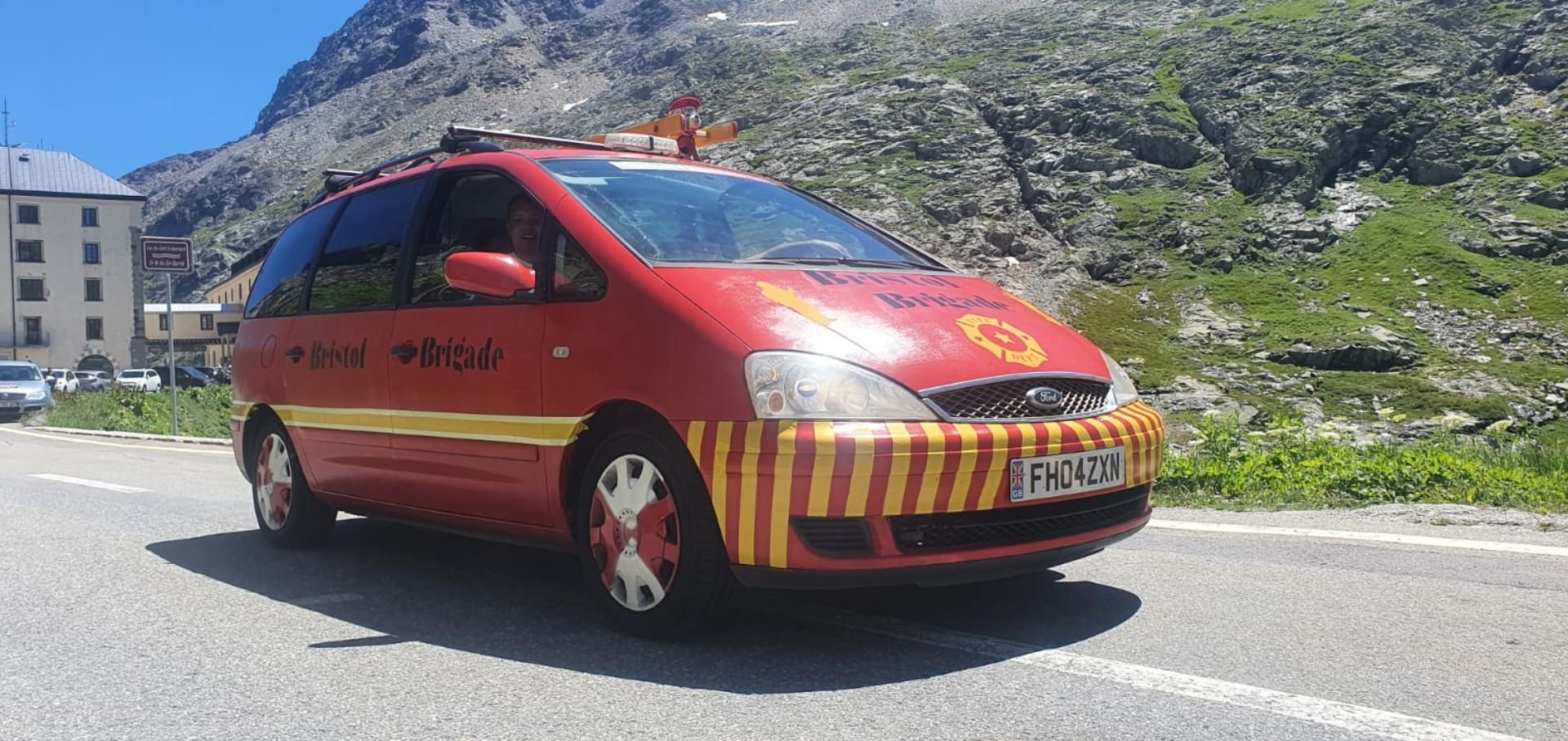 Red vehicle with ambulance markings and 'Bristol Brigade' written on the side, driving along a mountainous road with rocky terrain and green vegetation in the background.