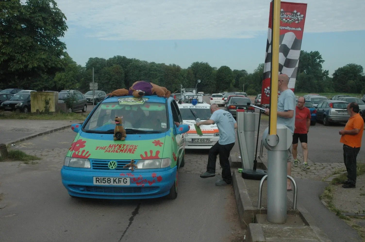 A colorful Volkswagen car decorated with flowers and the words 'The Mystery Machine' on the front, resembling the van from Scooby-Doo, parked in a parking lot with several people around it. One person is working on the car, and a large Scooby-Doo plu