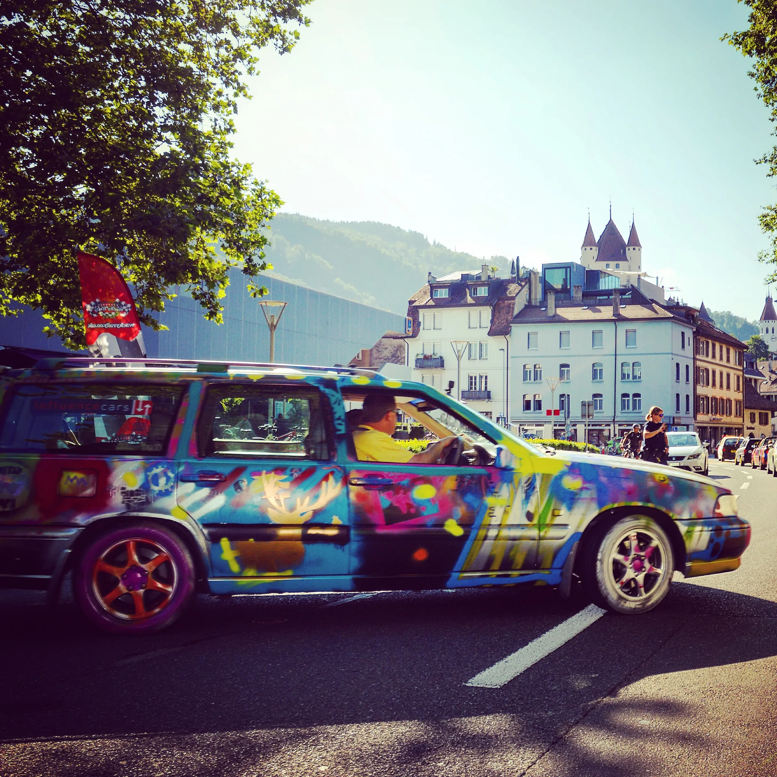 Colorfully painted car with graffiti-style art on a city street, with people walking and buildings in the background.