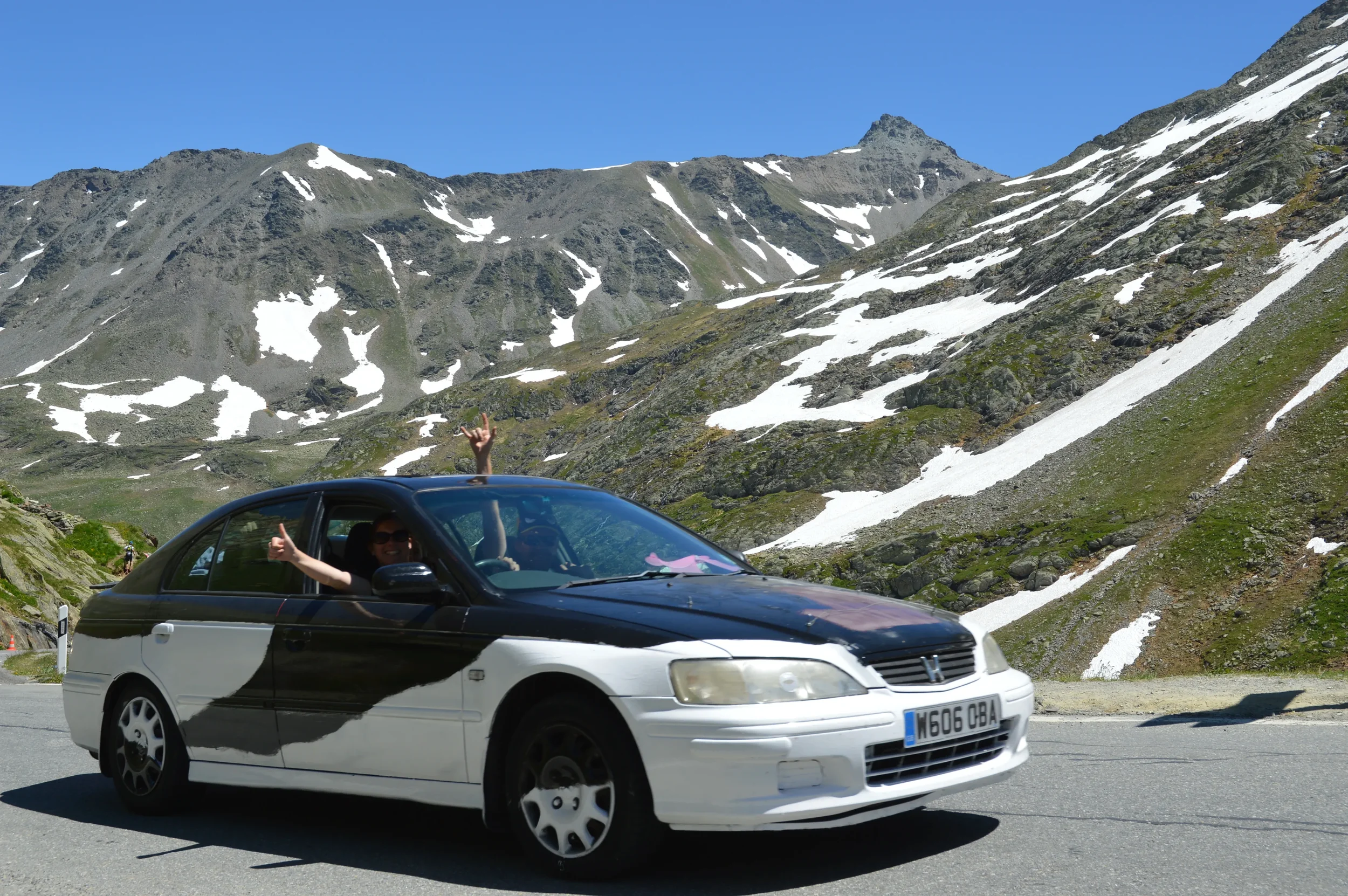 A person in a black and white Honda car making a peace sign with one hand and a thumbs-up with the other, driving on a mountain road with snow-capped peaks and a clear blue sky in the background.