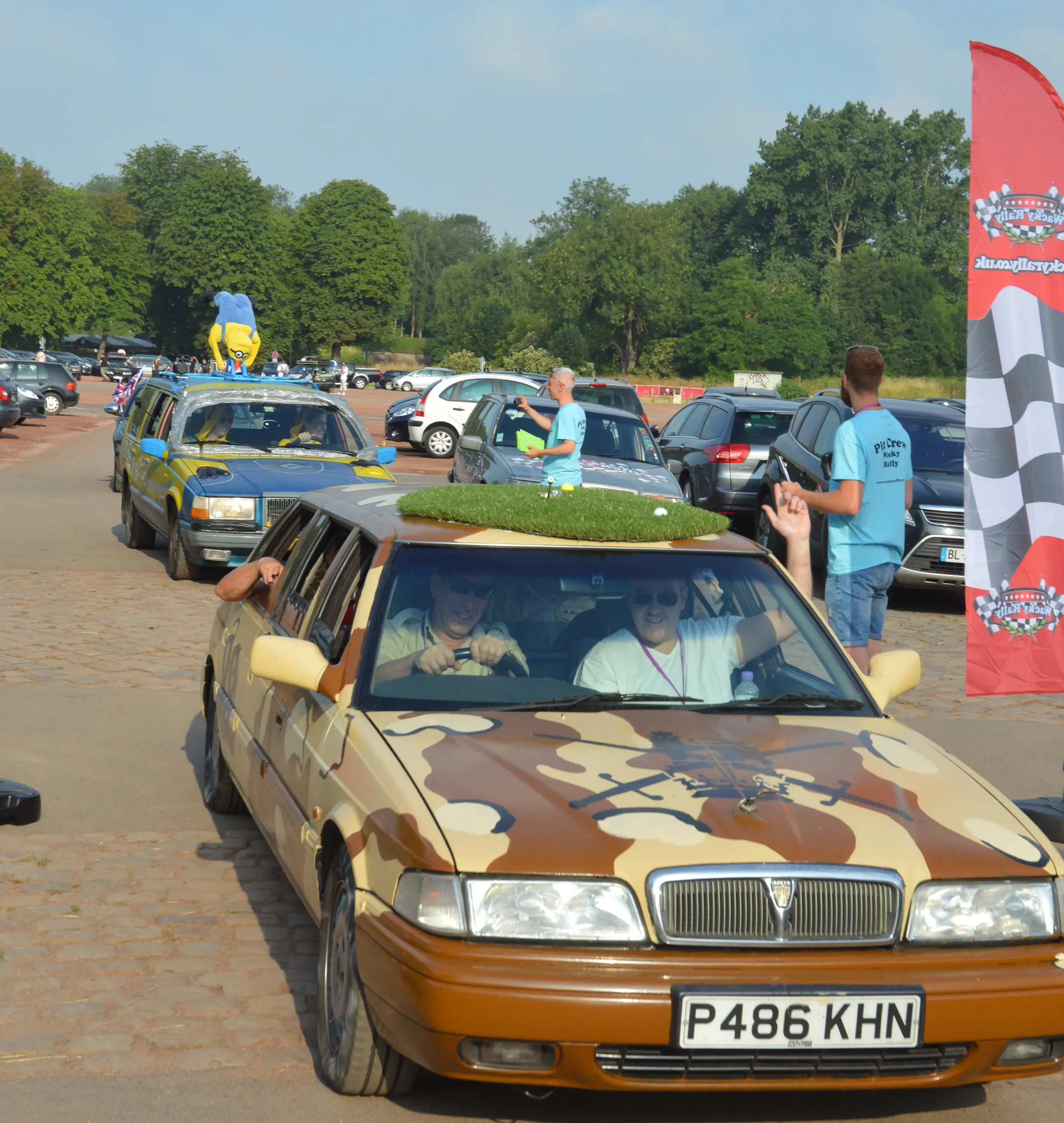 A vintage parade with decorated cars, including a camouflage-patterned vehicle with a grass-covered roof and a Trimoon mascot in the background. Several people are participating and watching, some taking photos.