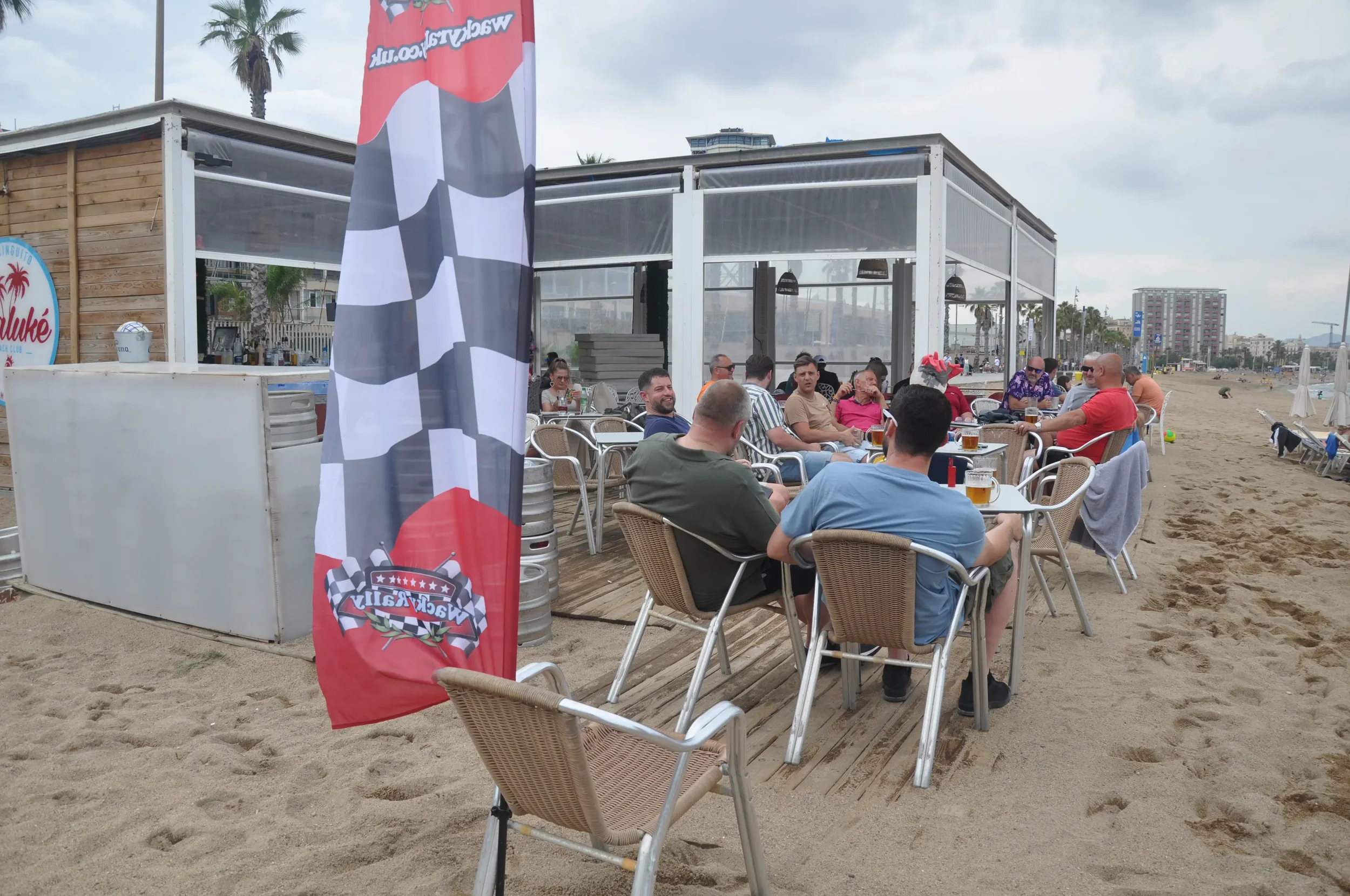 A group of people dining outdoors on a beachside patio, with tables, chairs, and a flag with racing-themed graphics.