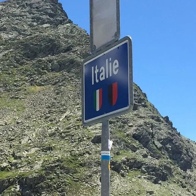 A road sign indicating entry into Italy, with the word 'Italie' and the Italian flag, set against a mountainous landscape with a clear blue sky.