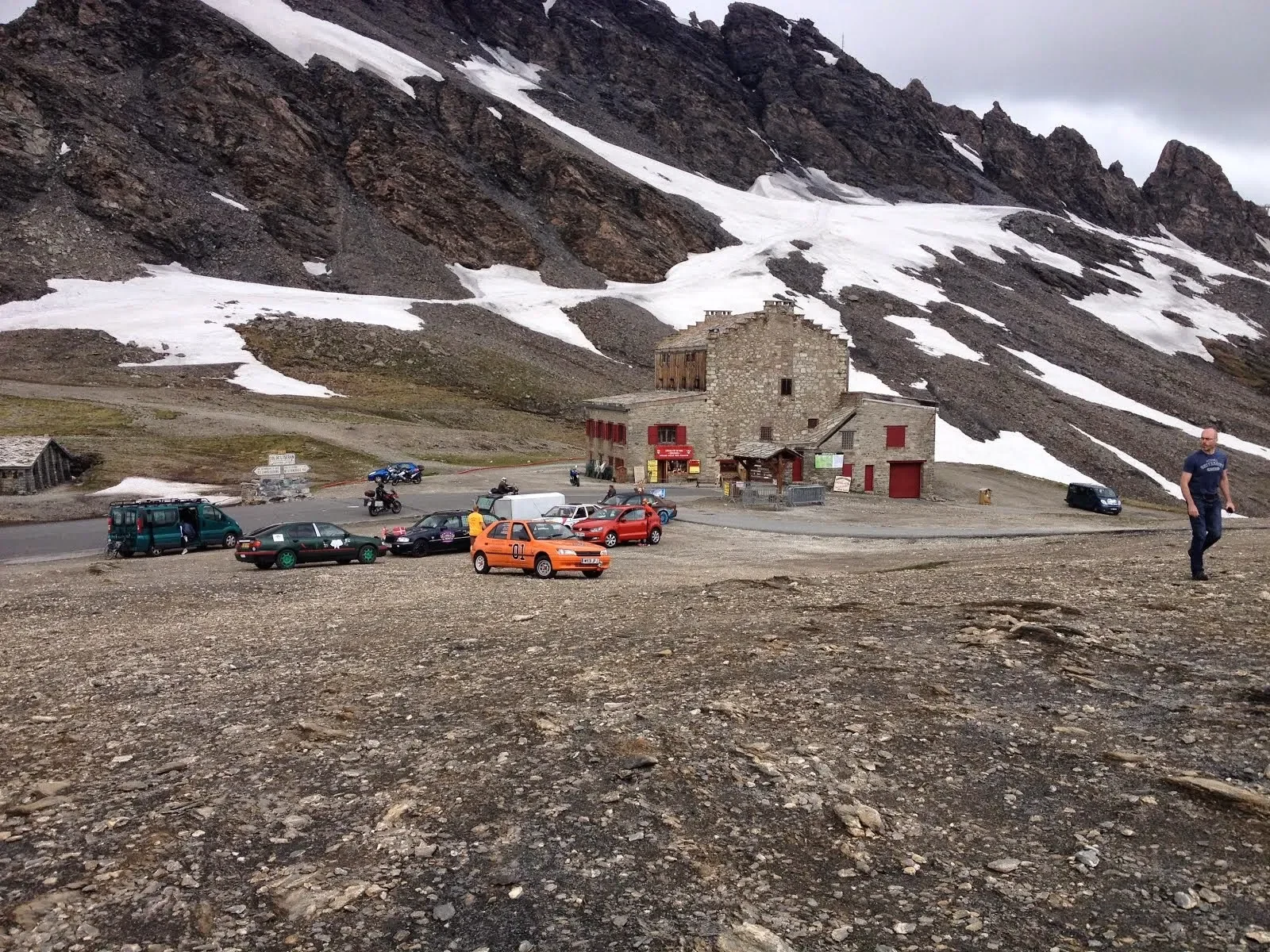 A mountainous area with patches of snow and several parked cars near a stone building. A man is walking on the rocky ground in the foreground.