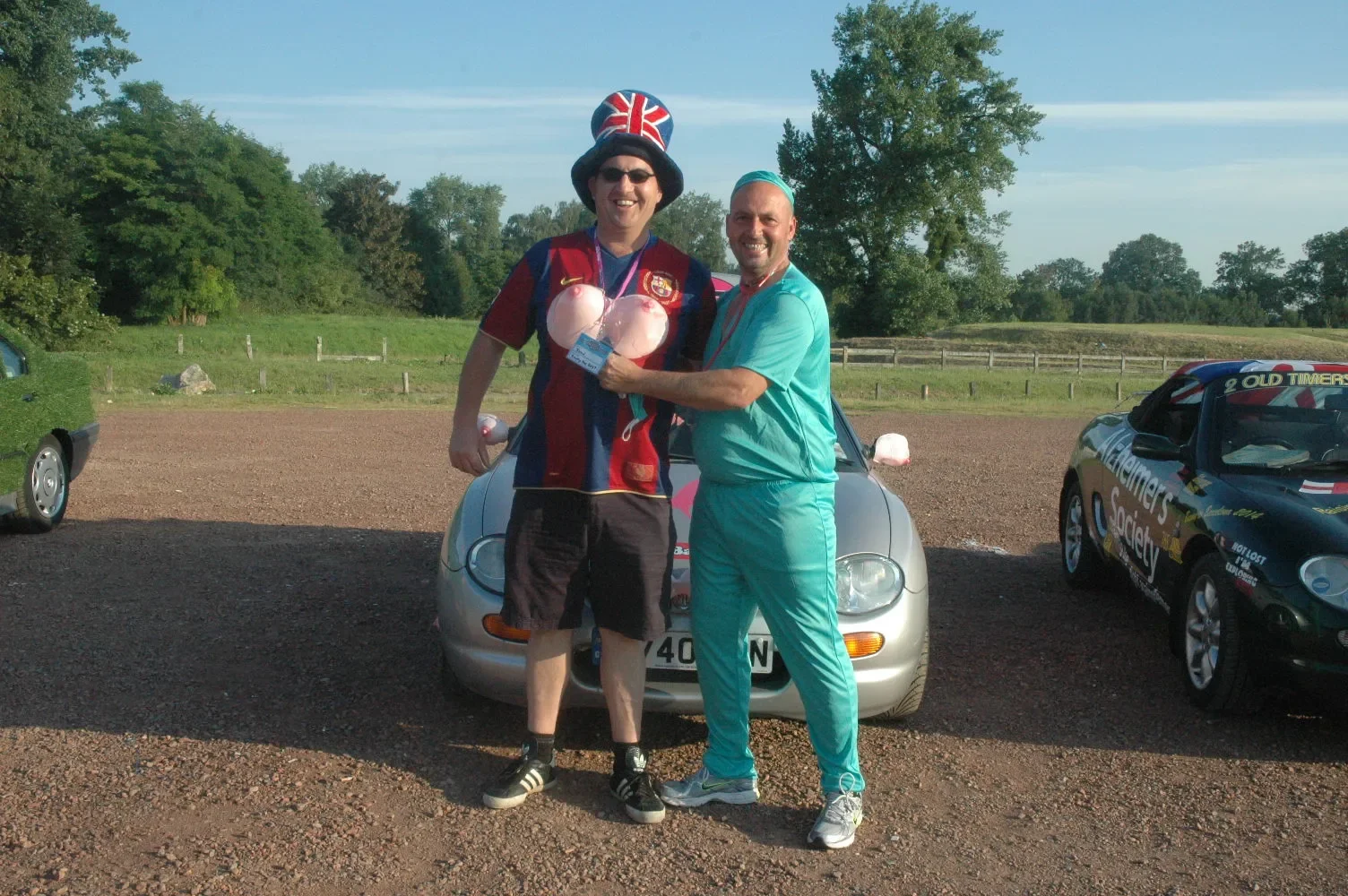 Two men standing in a parking lot, smiling and embracing each other. One man is wearing a sports jersey, shorts, sunglasses, and a hat with the British flag on top. The other man is dressed in light blue scrubs. They are holding pink balloons. There 