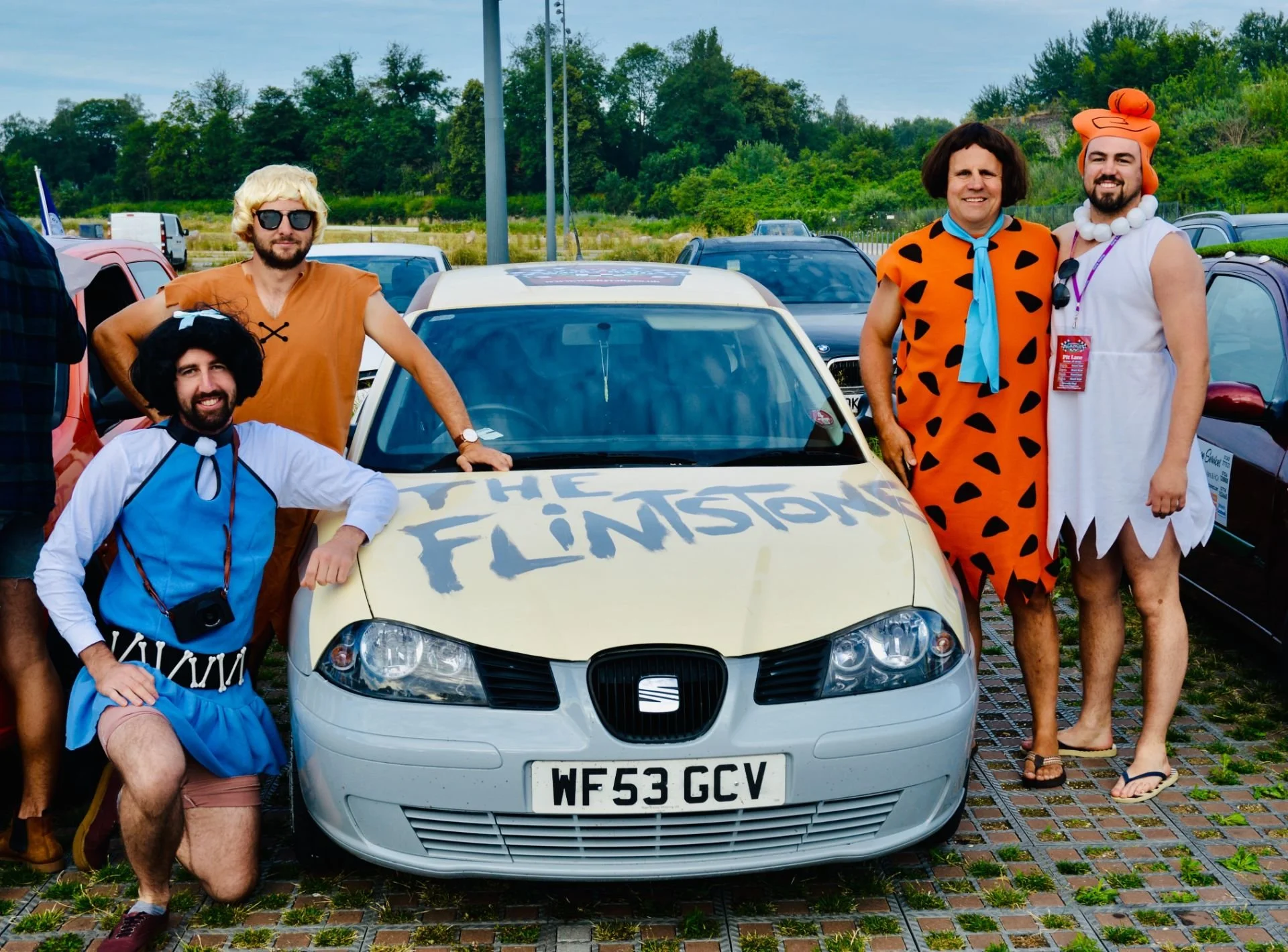 Group of five men dressed as characters from The Flintstones standing around a white car with "The Flintstones" written on the hood. One man is kneeling in front, another is leaning on the hood, and three are standing behind the car. They are in a pa