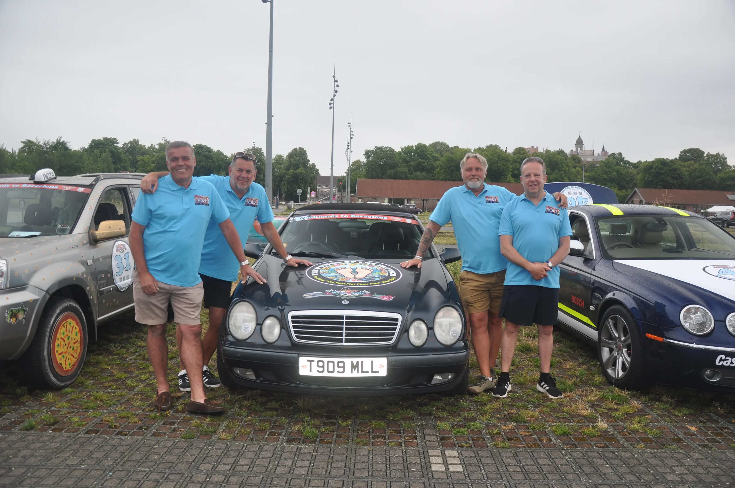 Five men in matching blue shirts standing next to decorated cars in an outdoor parking lot.