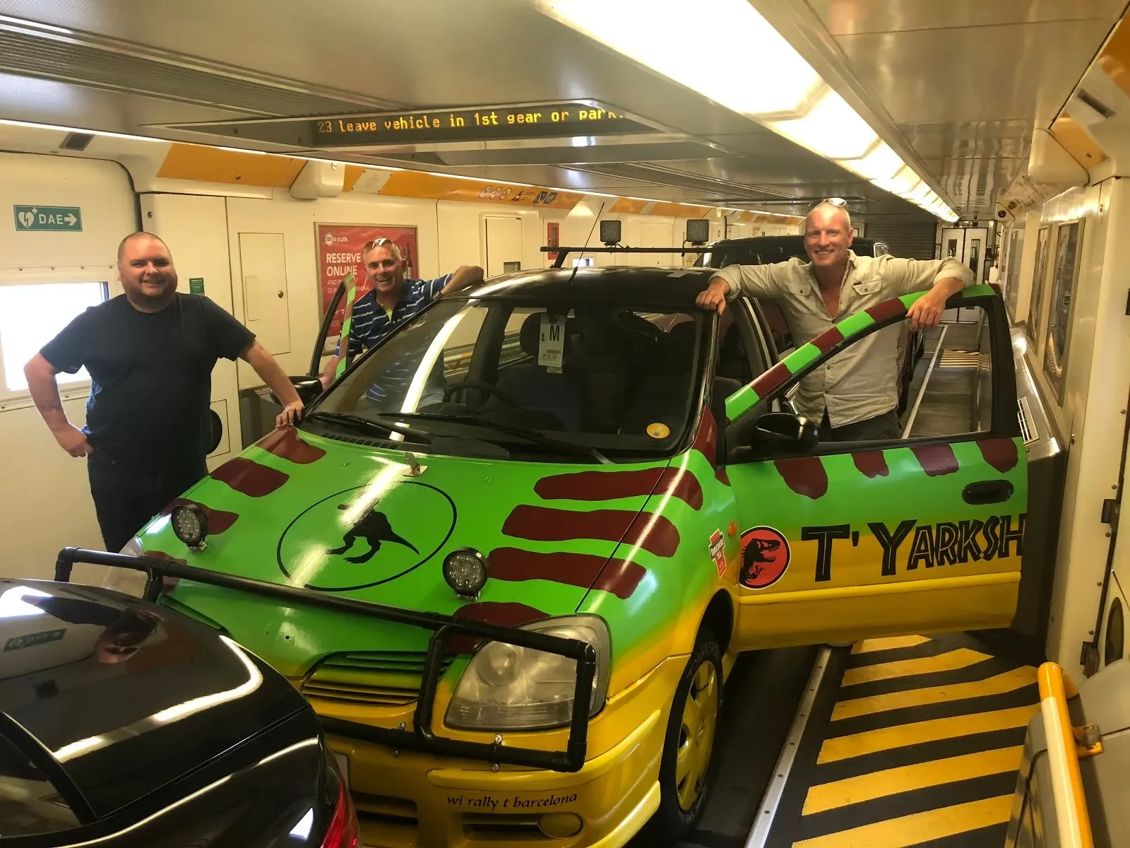 Three men standing around a small colorful car inside a parking garage. The car has a green and yellow paint job with red tiger stripe patterns and a logo featuring a dinosaur on the side that reads 'T YARKISH.'