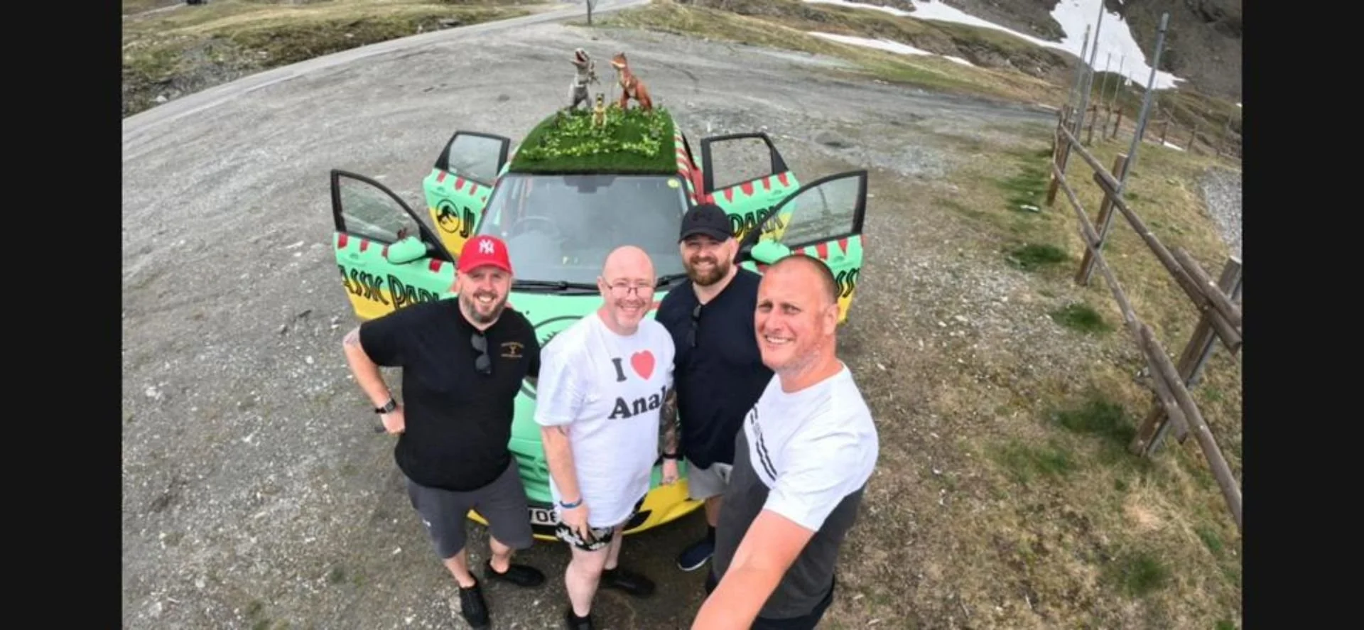 Four men taking a selfie in front of a colorful car with open doors and a roof decorated with dog figures. The men are smiling and standing on a gravel area with a wooden fence on the right side.