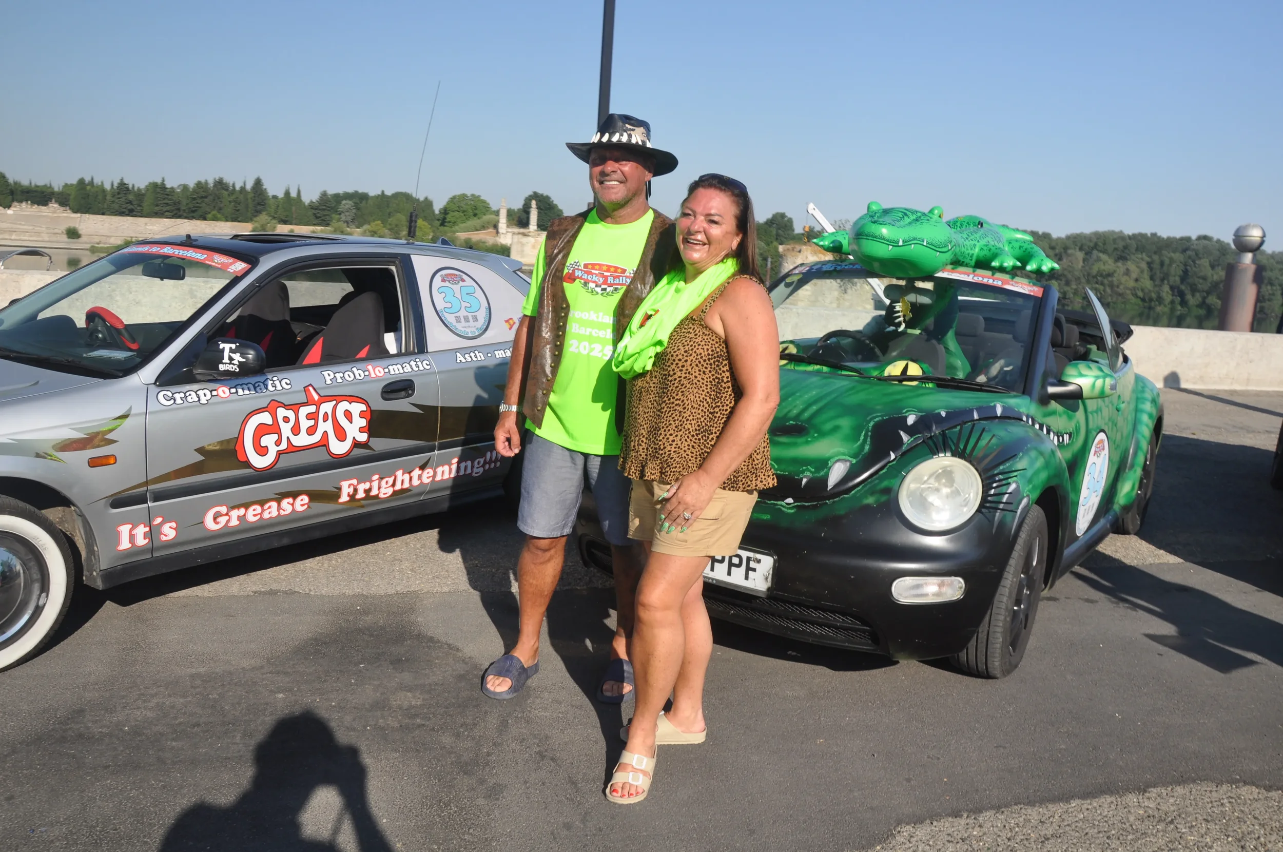 A man and woman standing in front of cars decorated with 'Grease' theme, featuring a crocodile puppet on one car, on a sunny day