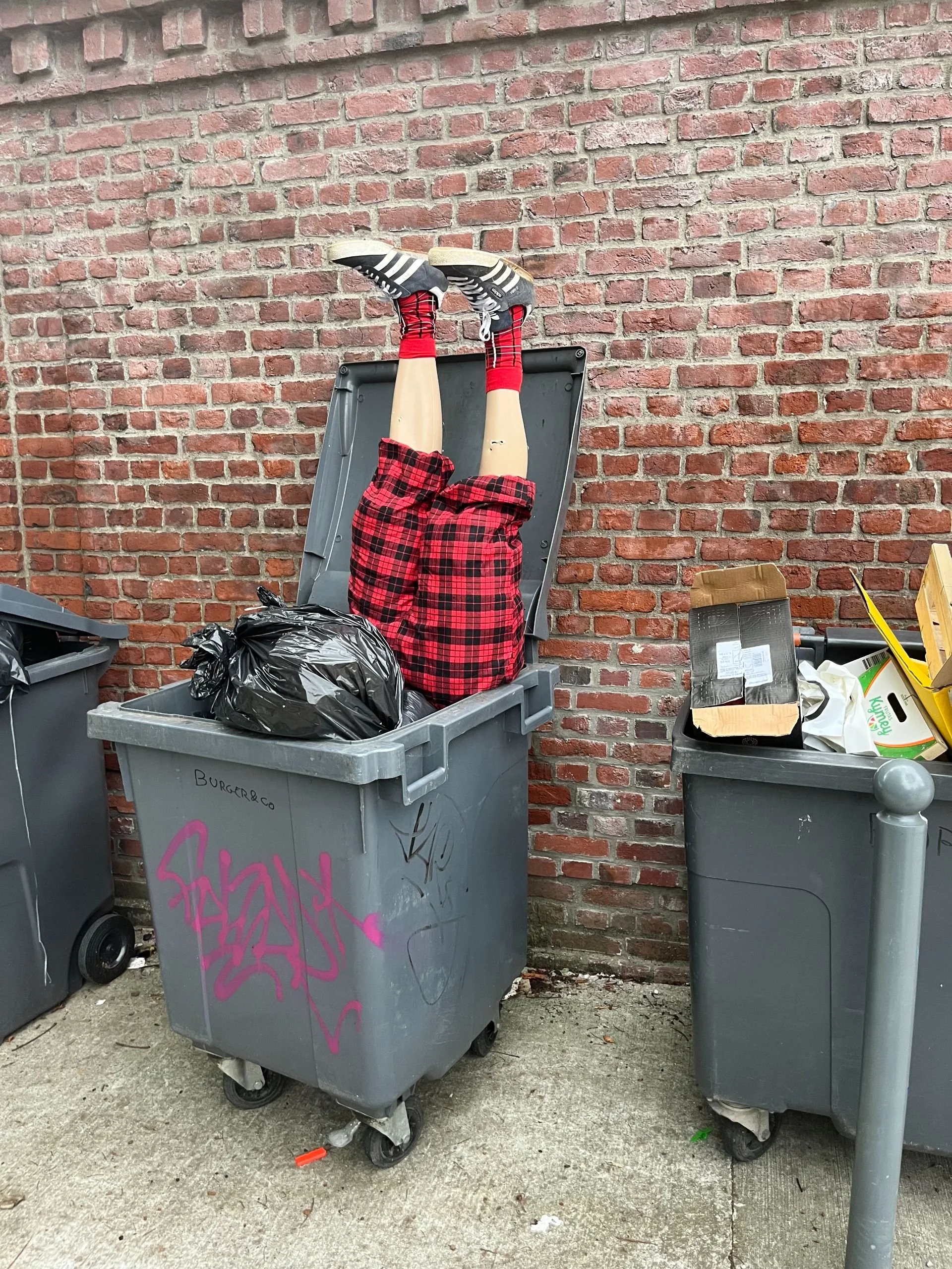 An overflowing trash bin with legs and shoes sticking out, next to other trash bins and a brick wall.