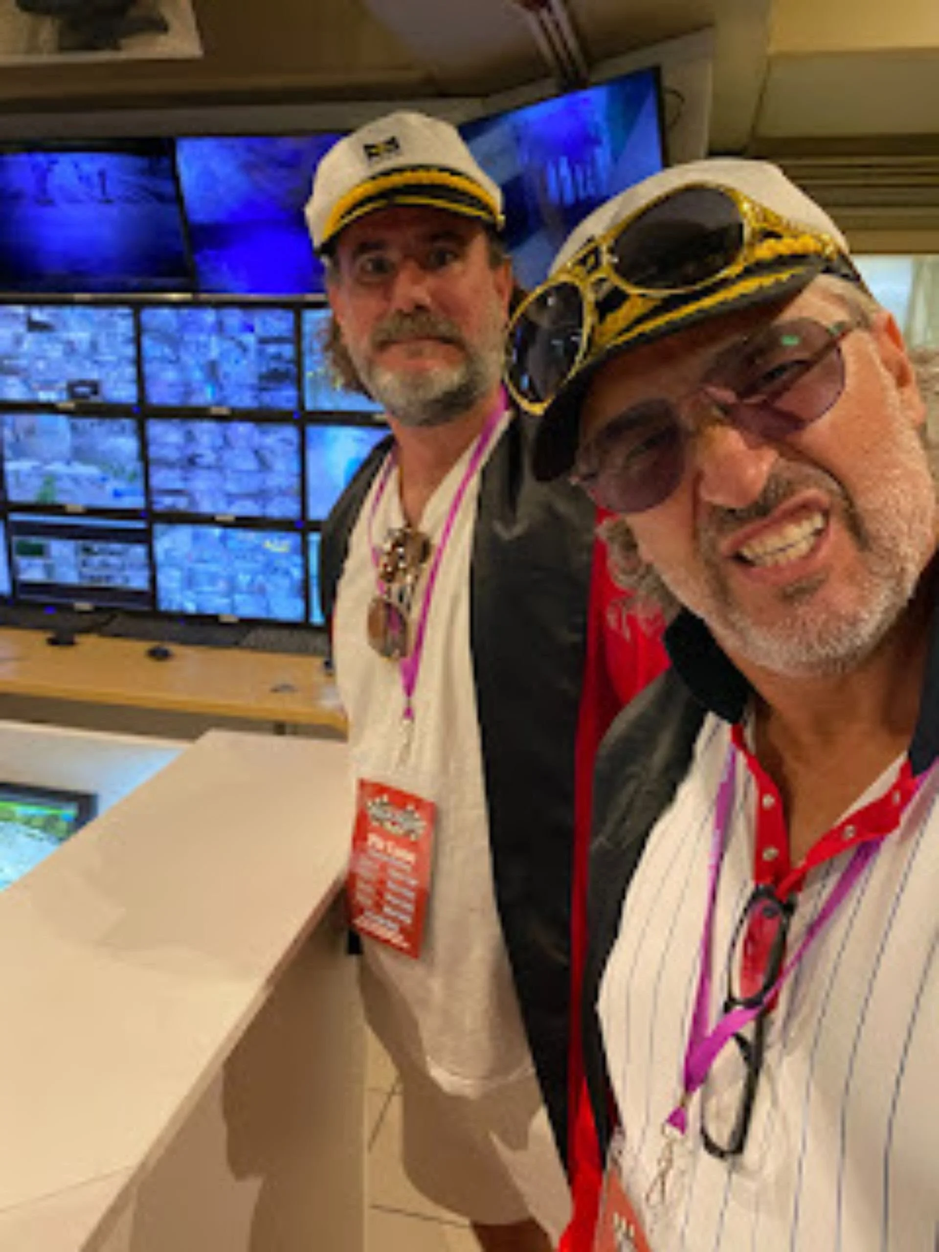 Two men wearing captain hats and sunglasses taking a selfie in front of a wall of security camera monitors.