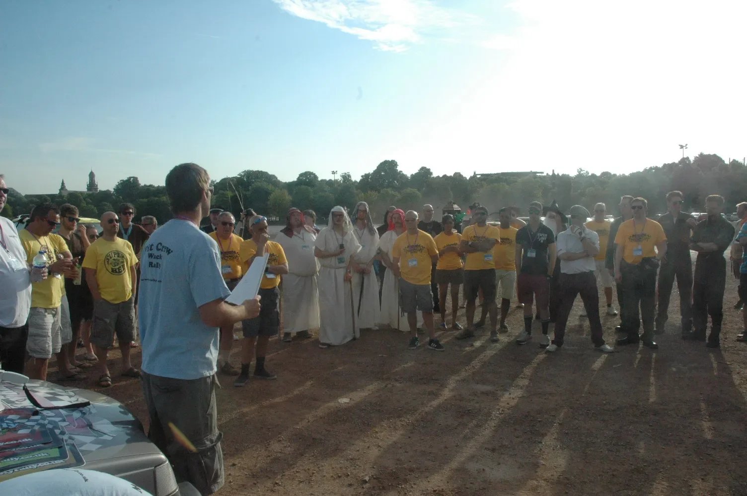 A man in a light blue shirt appears to be addressing a large group of people outdoors on a sunny day. The group consists of men and women, some wearing yellow shirts and others in white or darker clothing, standing on a dirt surface with trees and di